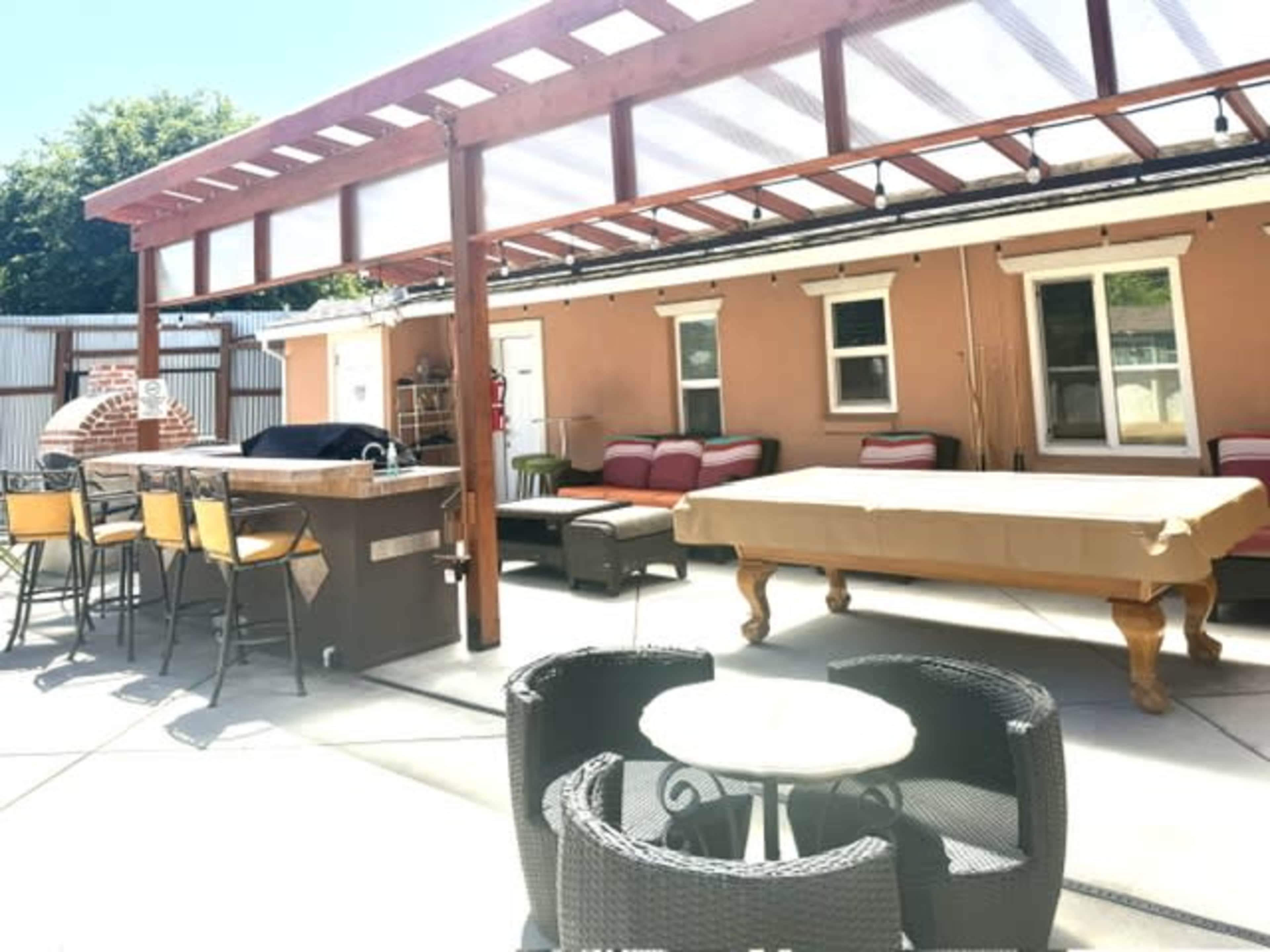 The image shows a patio area with outdoor seating, a bar counter, and a pool table under a pergola.