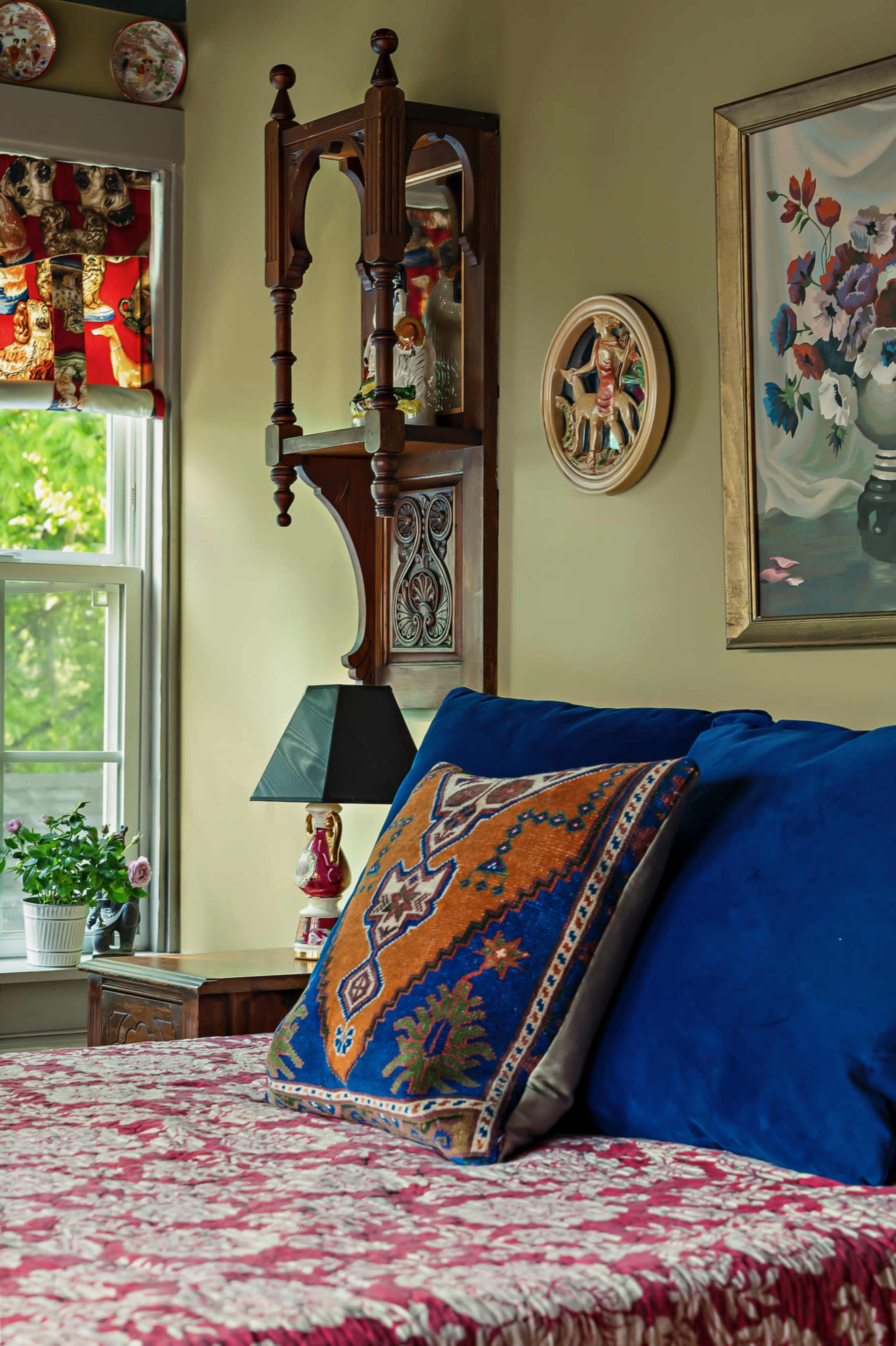 The image shows a cozy bedroom corner featuring a bed with decorative pillows, a wooden shelf with ornamental pieces, a table lamp, and a window with patterned curtains.