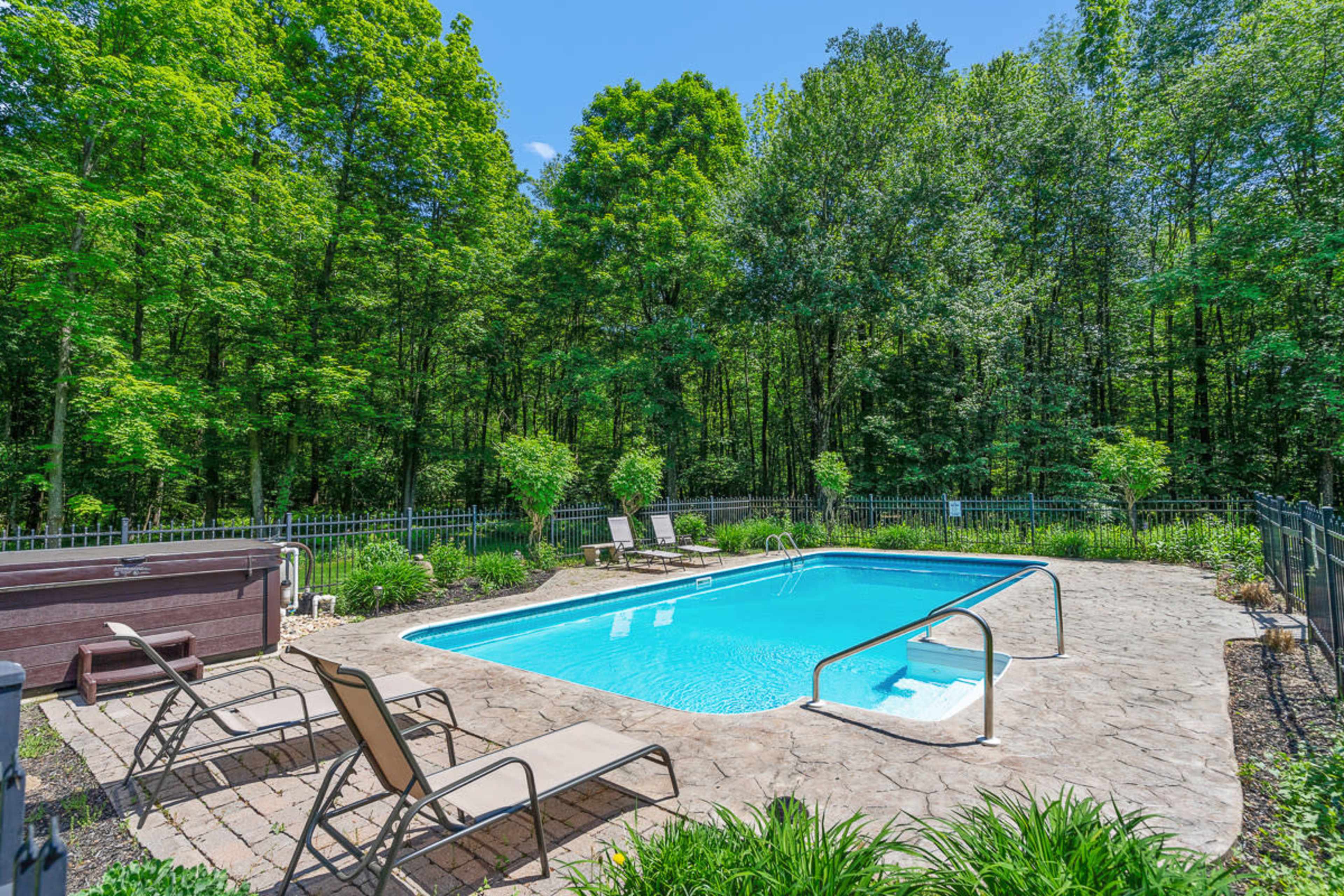 A rectangular swimming pool is surrounded by a stone patio and lounge chairs, nestled among lush green trees.