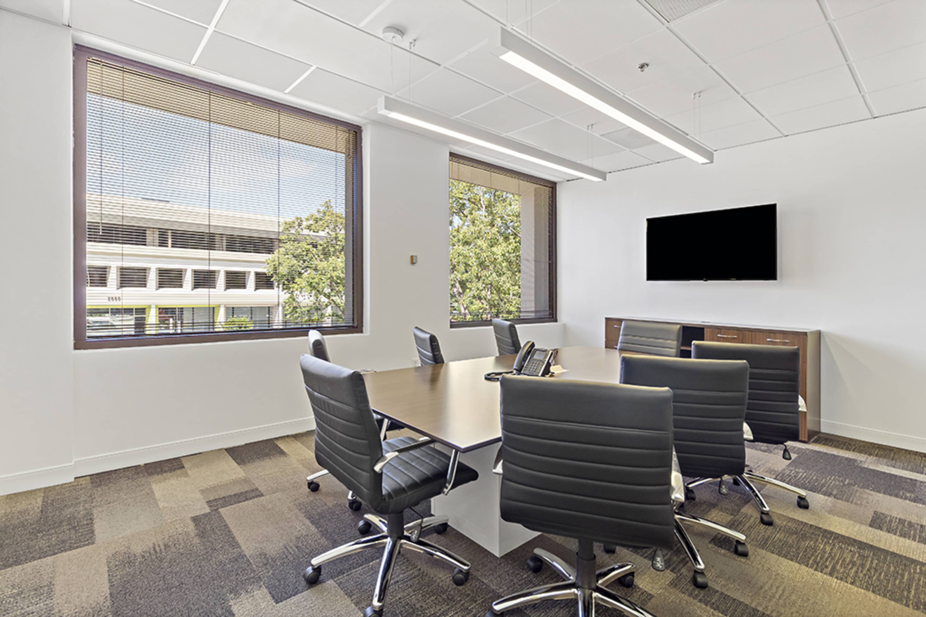 A conference room features a large table surrounded by black leather chairs, with windows offering a view of the outdoors and a wall-mounted television.