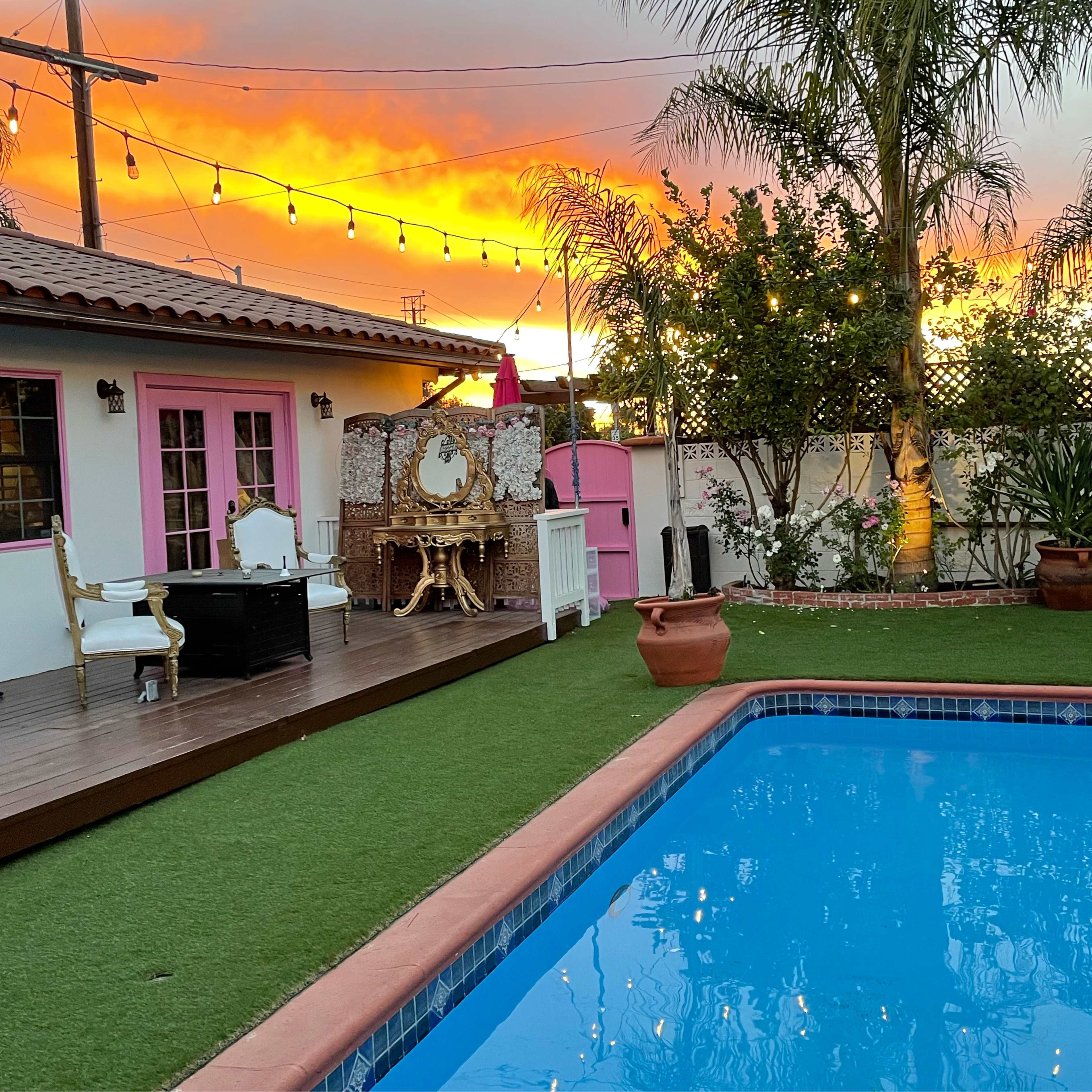 The scene shows a backyard with a swimming pool, a wooden deck, and a perfectly framed sunset in the background.