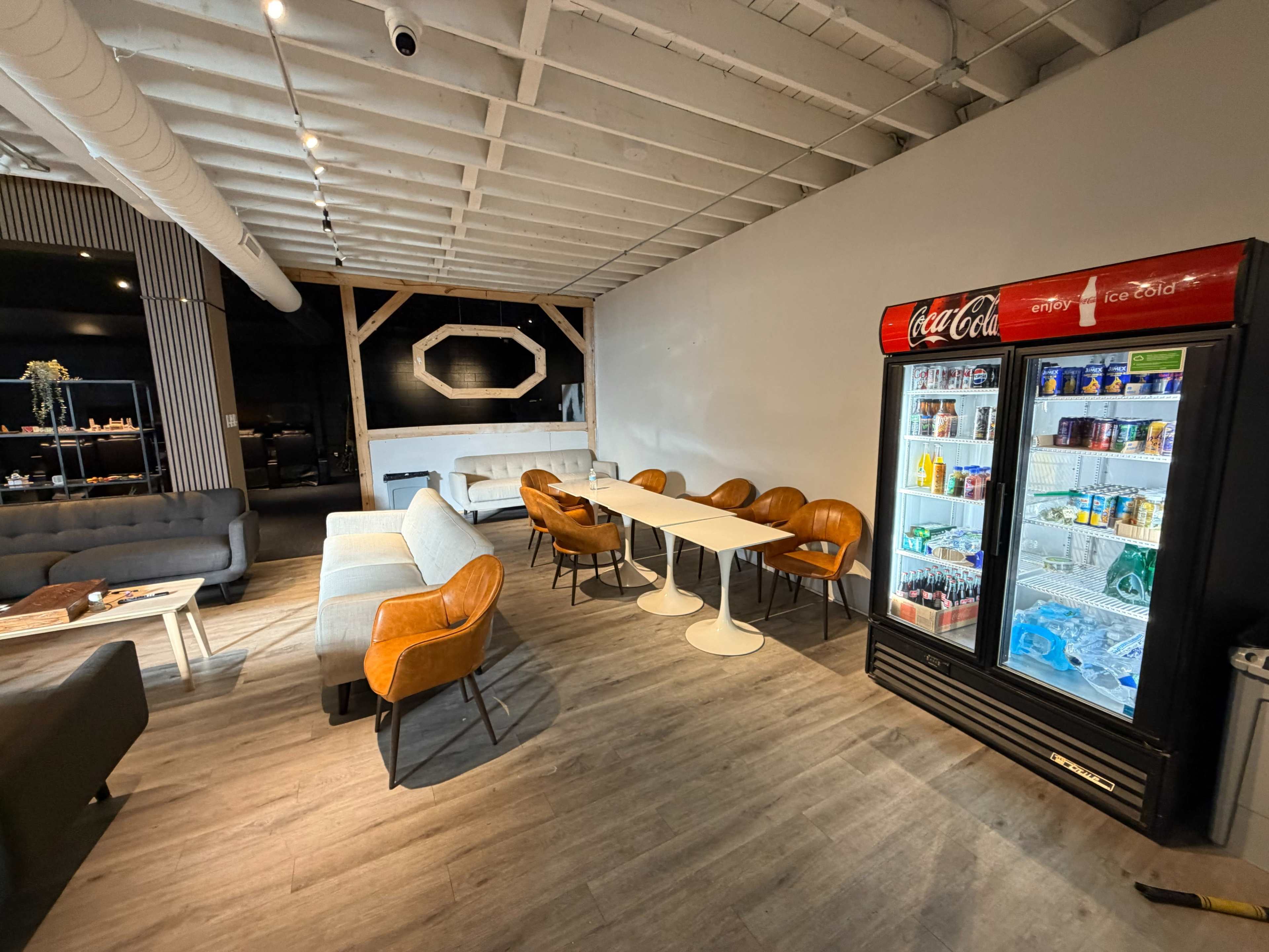 The image shows a modern indoor seating area featuring gray sofas, wooden tables, and a Coca-Cola vending machine next to a refrigerator displaying various drinks.