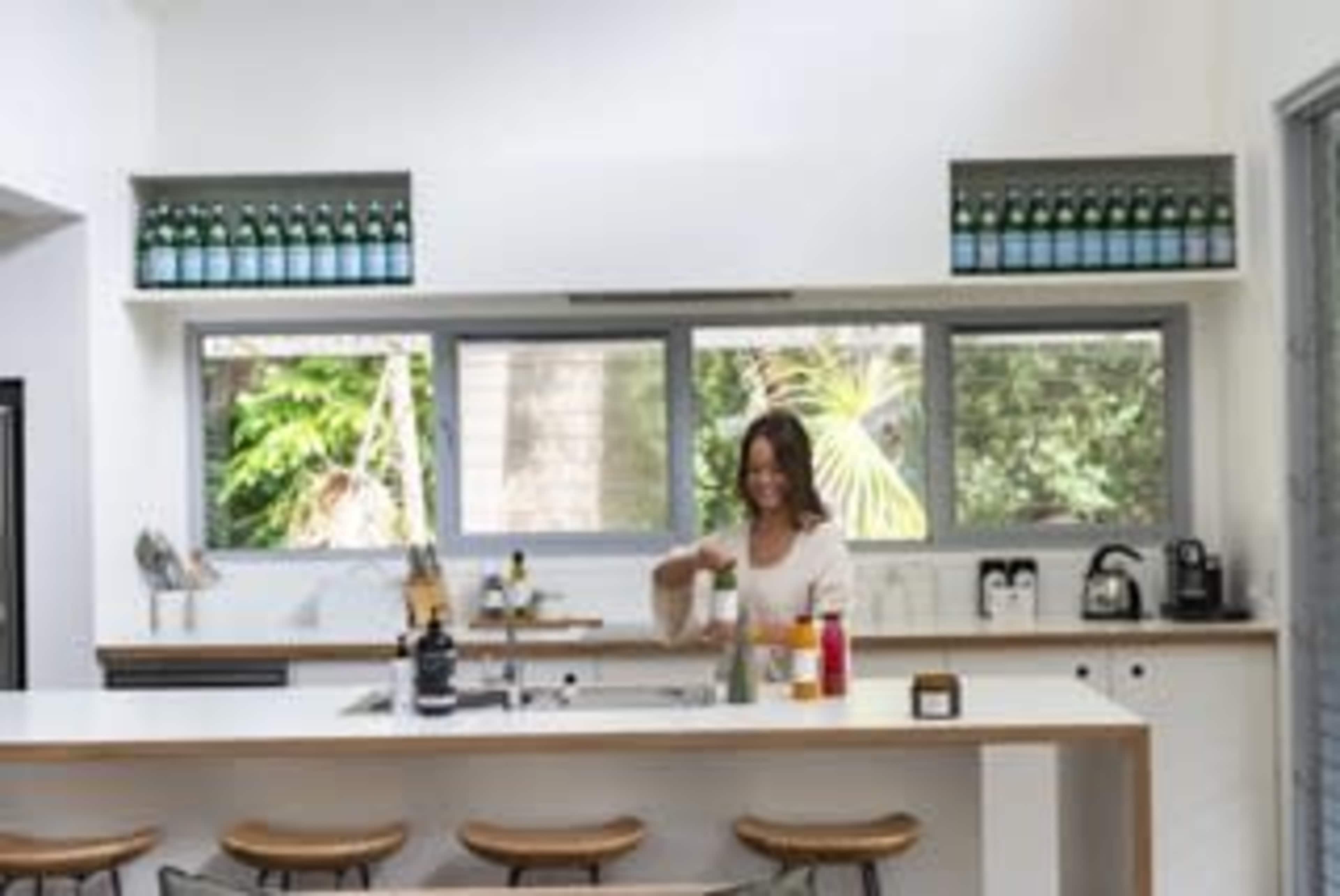 A woman prepares a drink at a modern kitchen island with bottles displayed on the shelves above and a view of greenery through large windows.