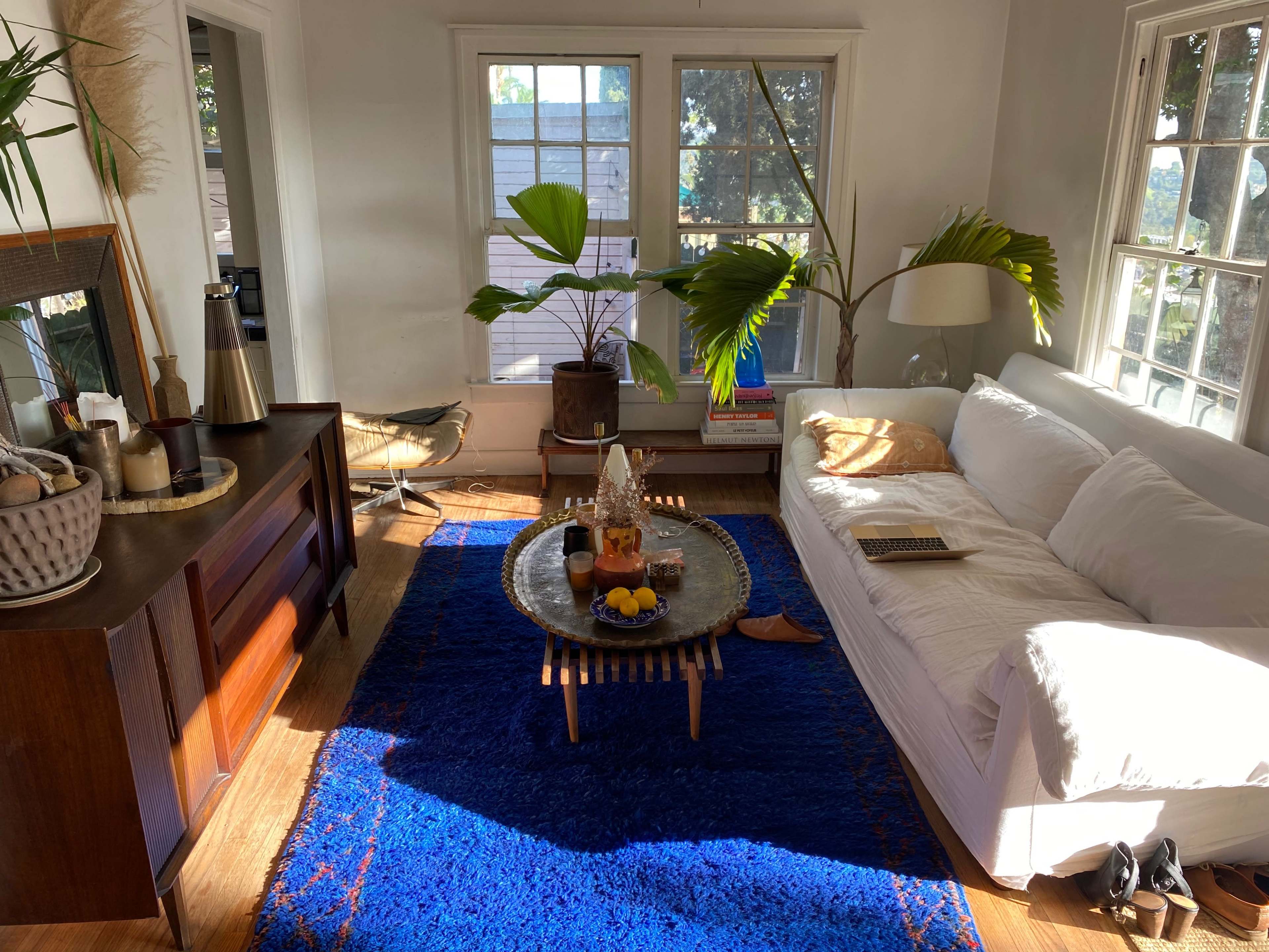 The scene features a living room with a white sofa, a wooden sideboard, a round coffee table adorned with fruit, and large windows allowing natural light to enter.