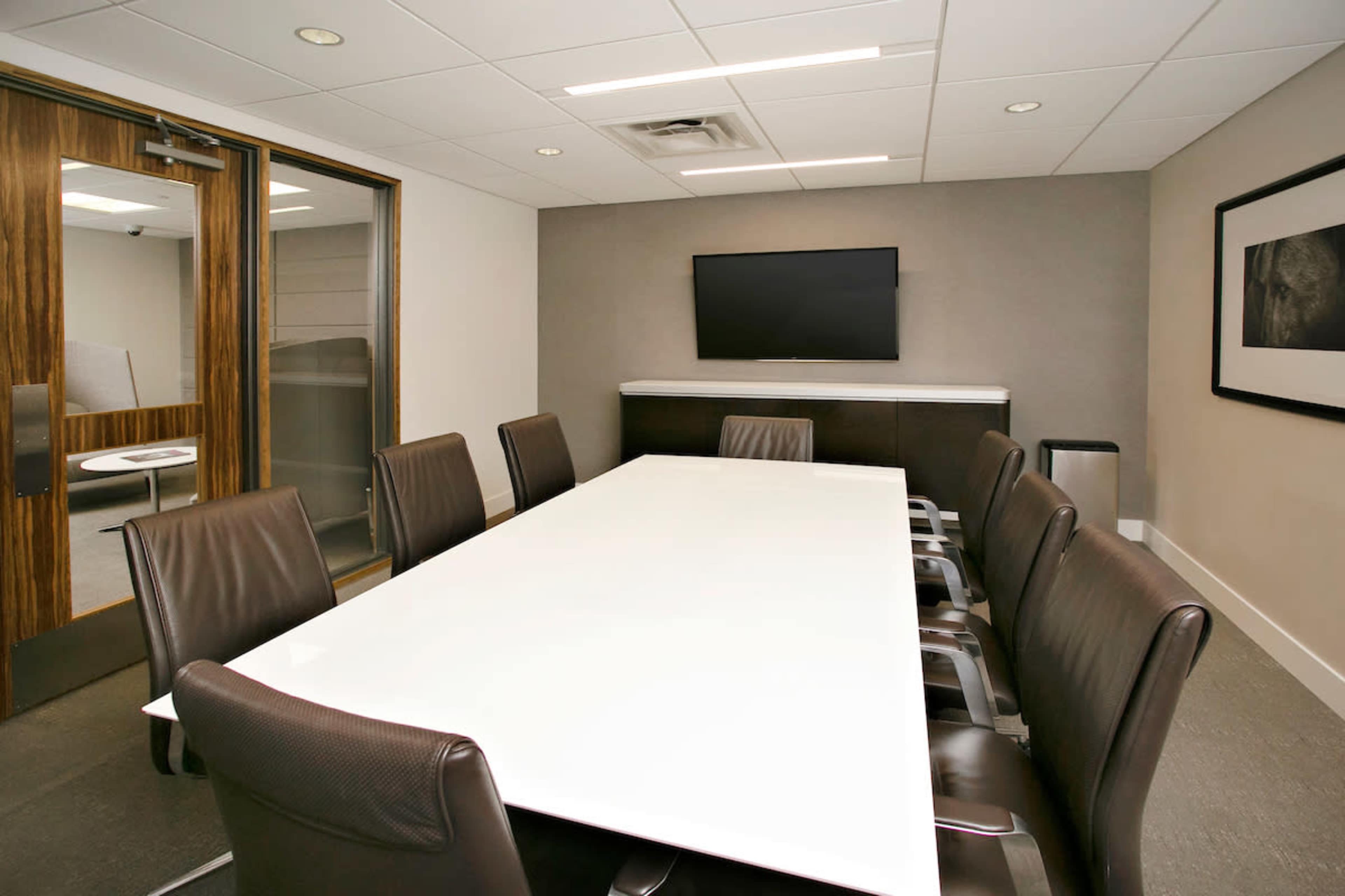 A modern conference room features a long white table surrounded by leather chairs and a wall-mounted television.