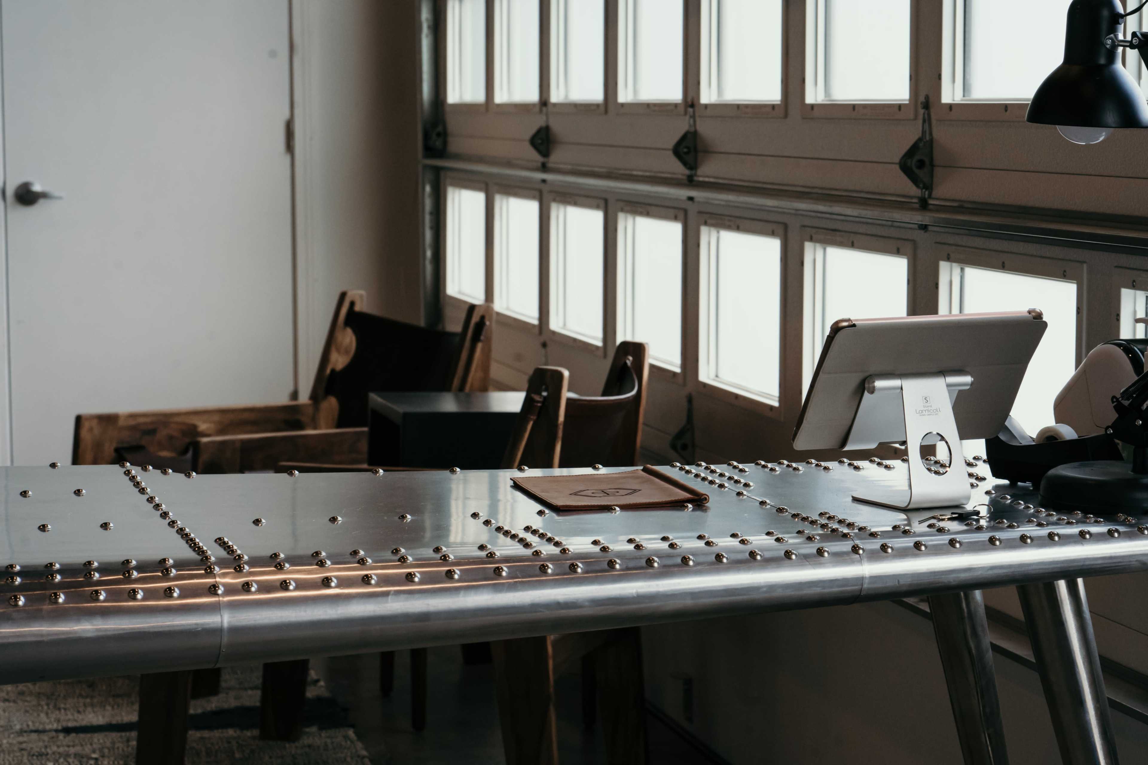 A metallic table with rivets and an iPad stand is situated in a room with large windows and wooden chairs.