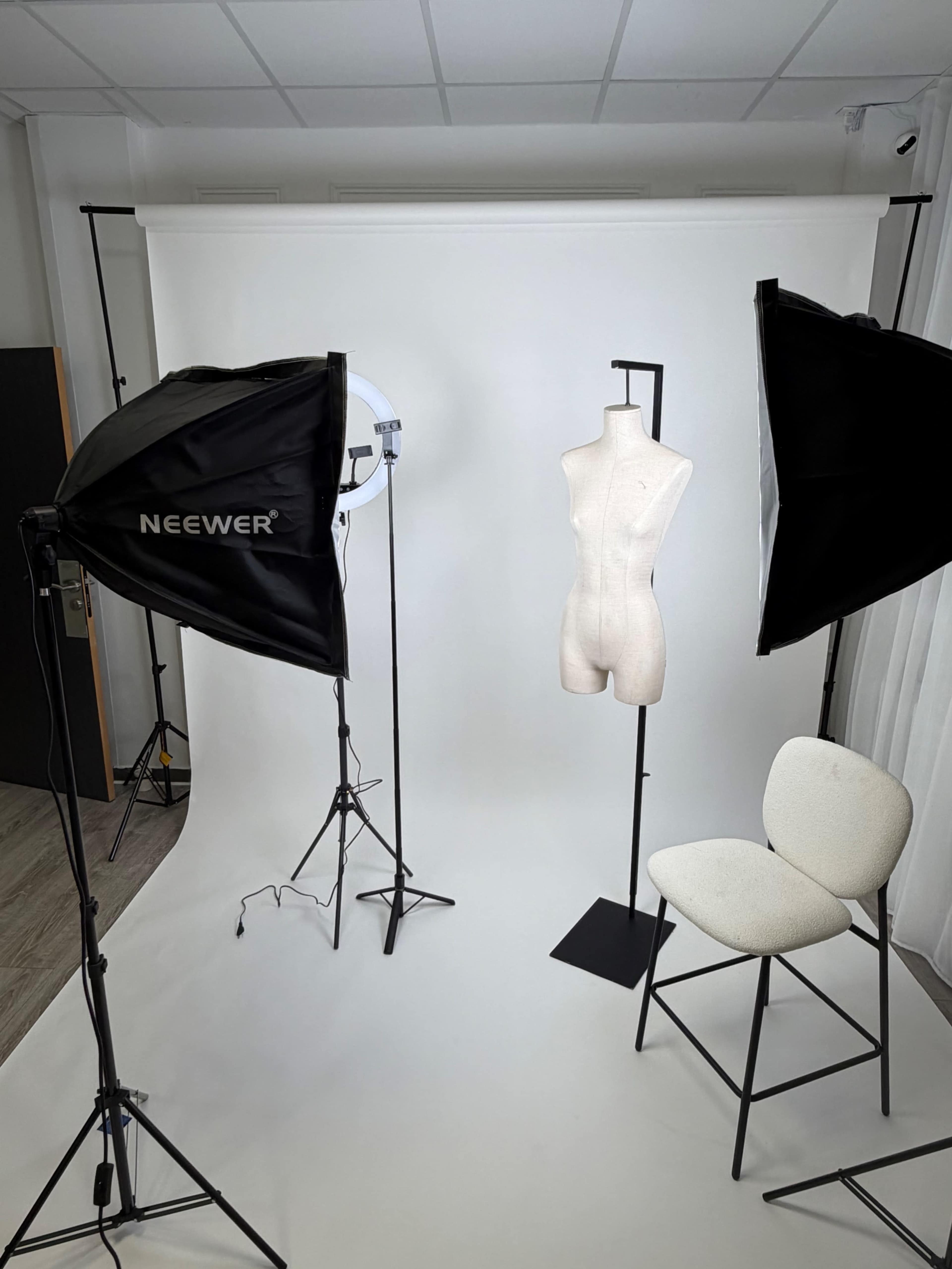 The image shows a photography studio setup featuring a dress form on a stand surrounded by two softbox lights and a chair, all against a plain white backdrop.