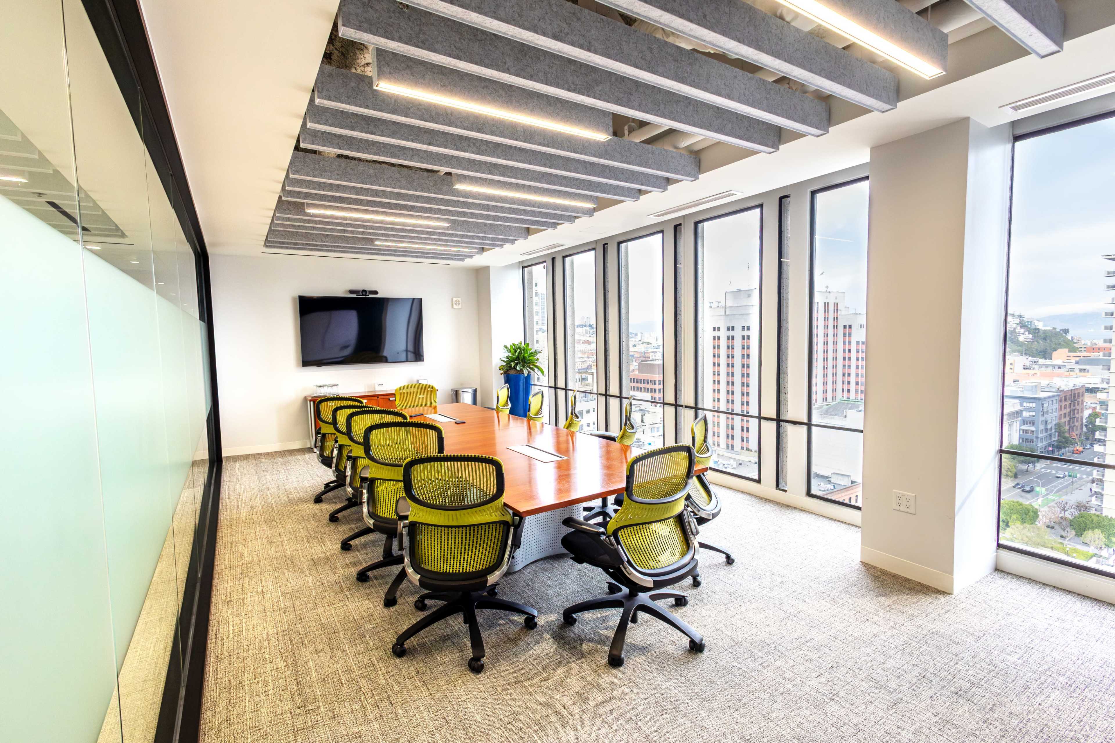 A modern conference room features a long wooden table surrounded by ergonomic chairs, with large windows offering a city view and a television mounted on the wall.