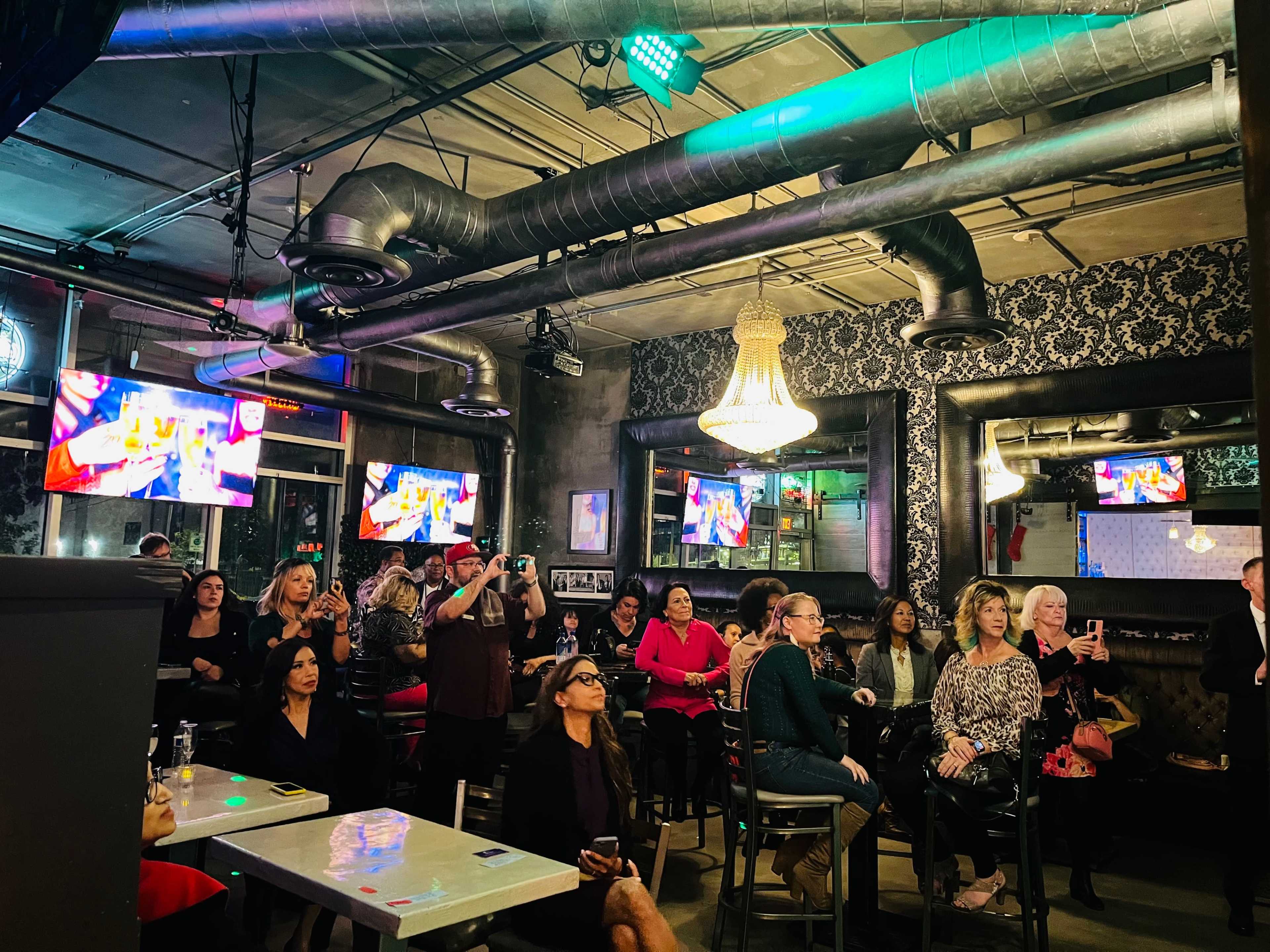 A crowd of people is seated in a bar, watching a presentation on multiple screens while others stand and take photos.
