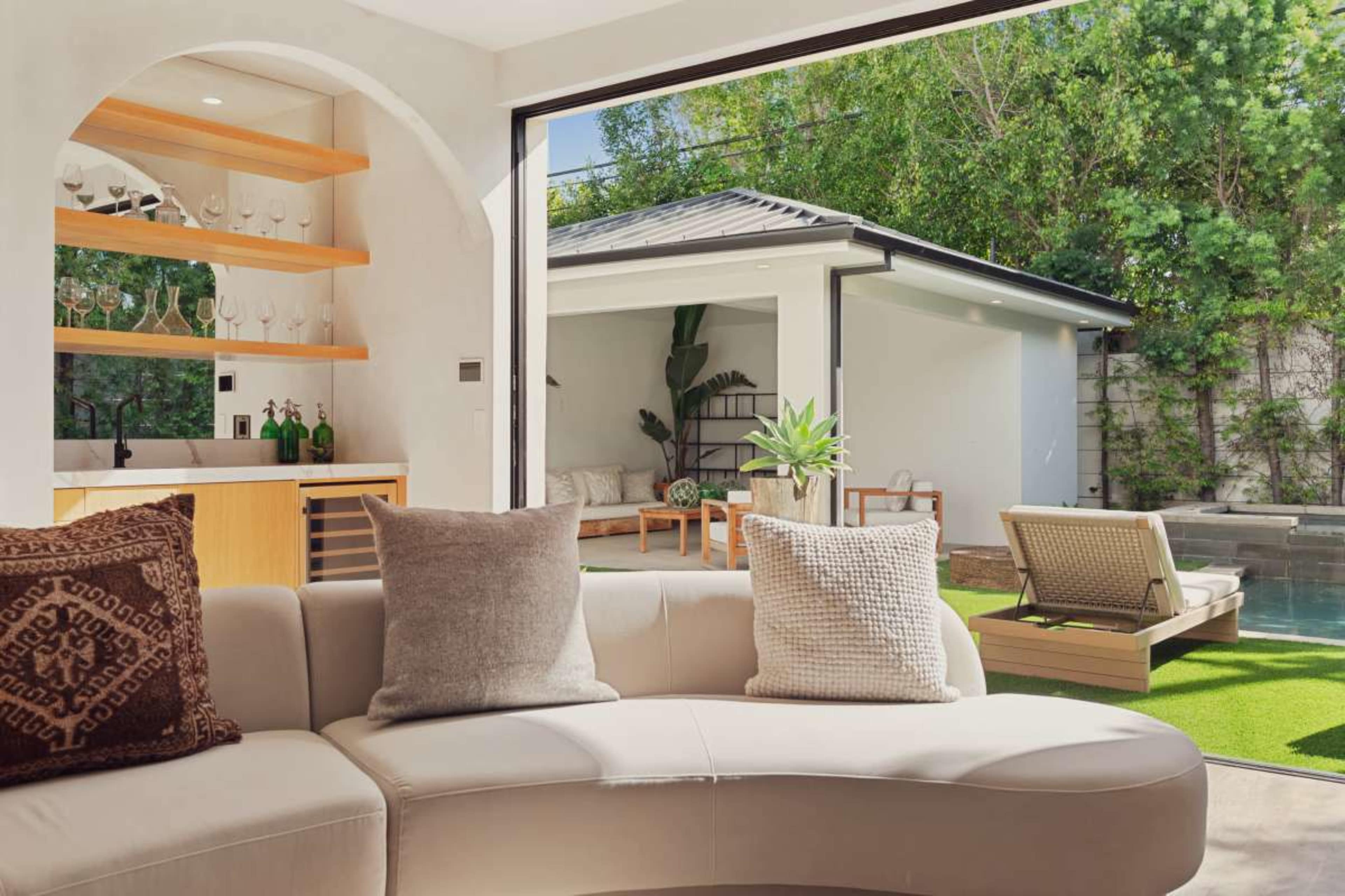 A modern living room features a curved sofa with decorative pillows, a bar area with glassware, and large sliding doors opening to a patio with seating and a pool.