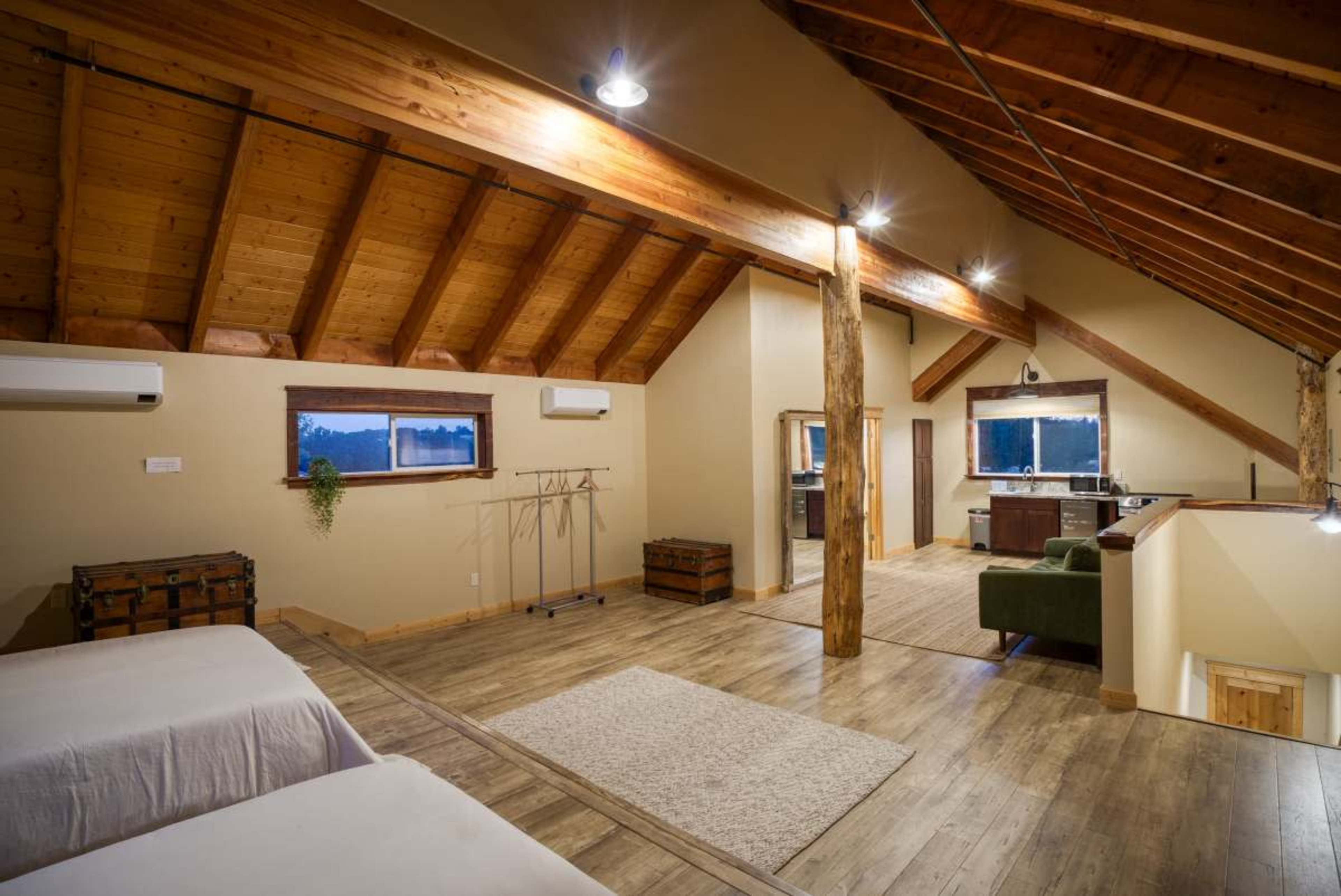 A spacious loft bedroom with wooden beams, a light wooden floor, two beds, and a seating area, alongside a kitchenette and large windows.