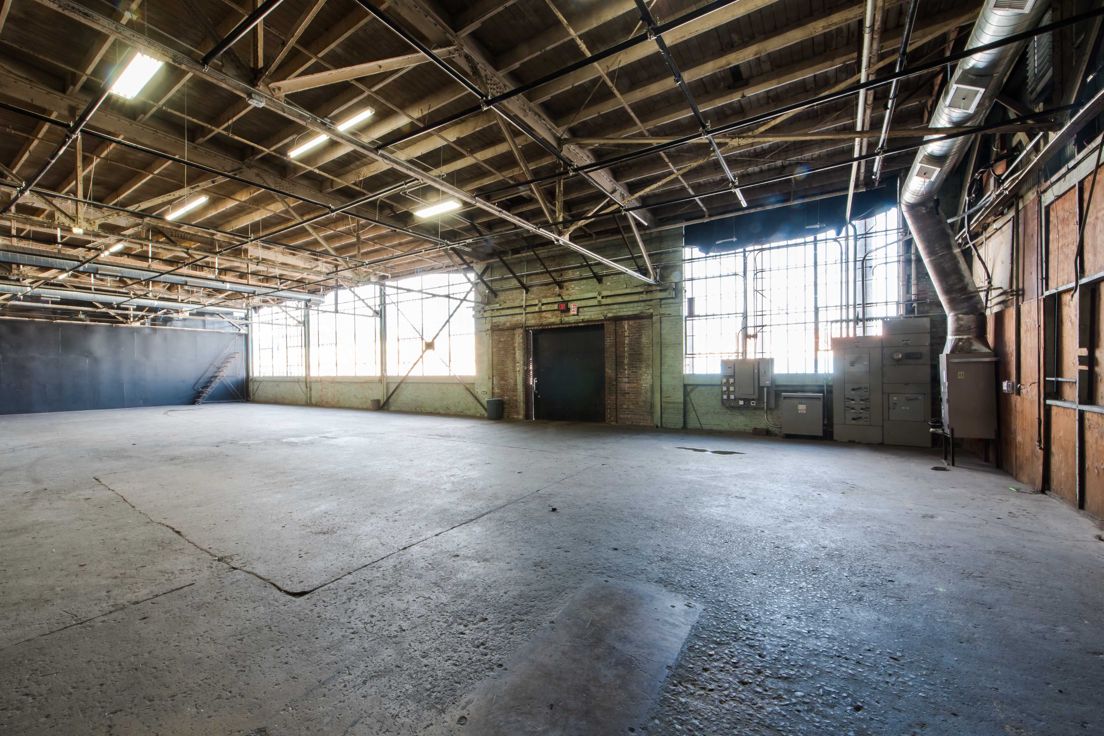 The image shows a large, empty industrial space with concrete flooring, exposed wooden beams, and large windows along one wall.
