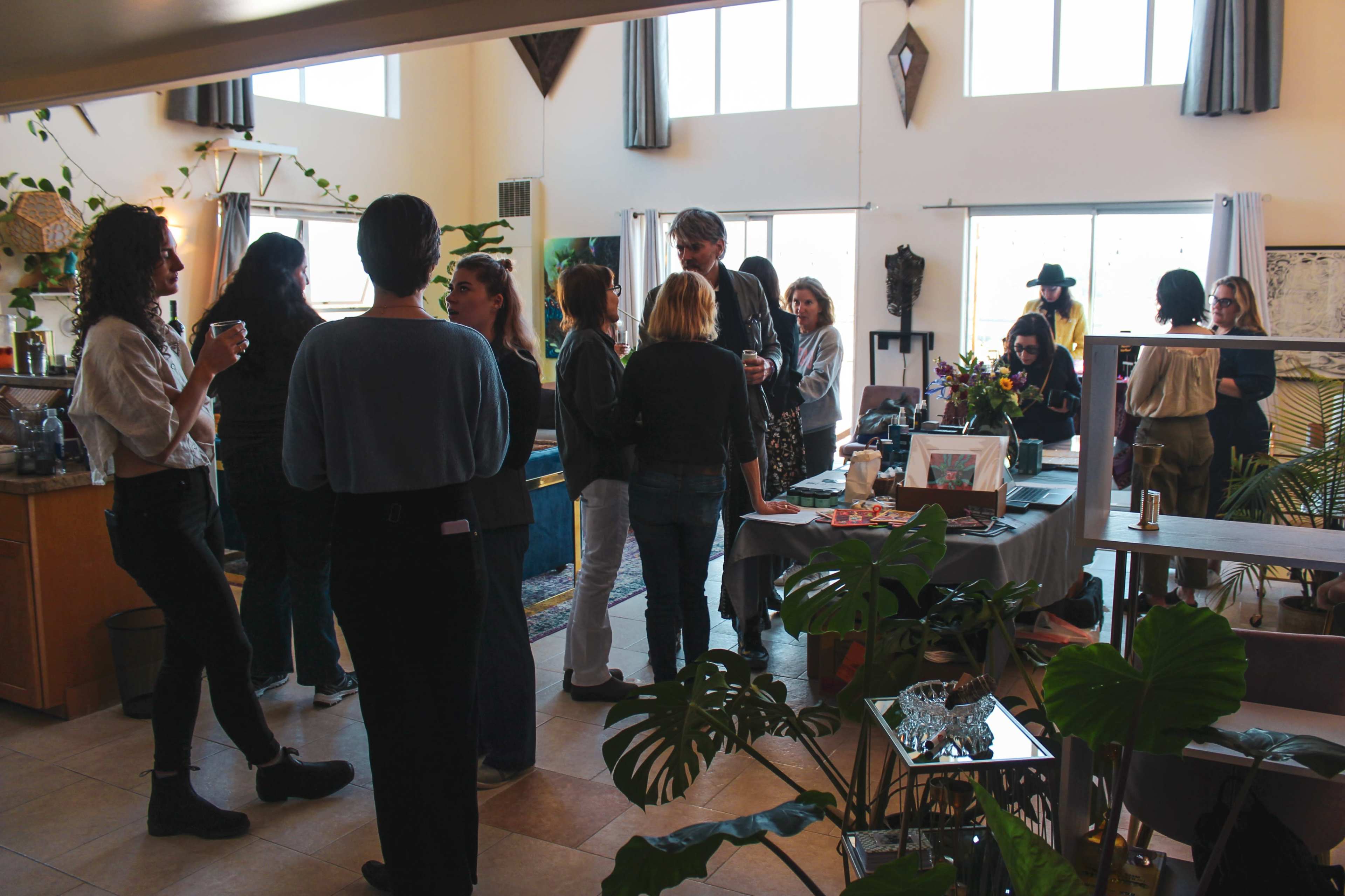 A diverse group of people mingles in a brightly lit room filled with plants, tables, and decorations during a social gathering.