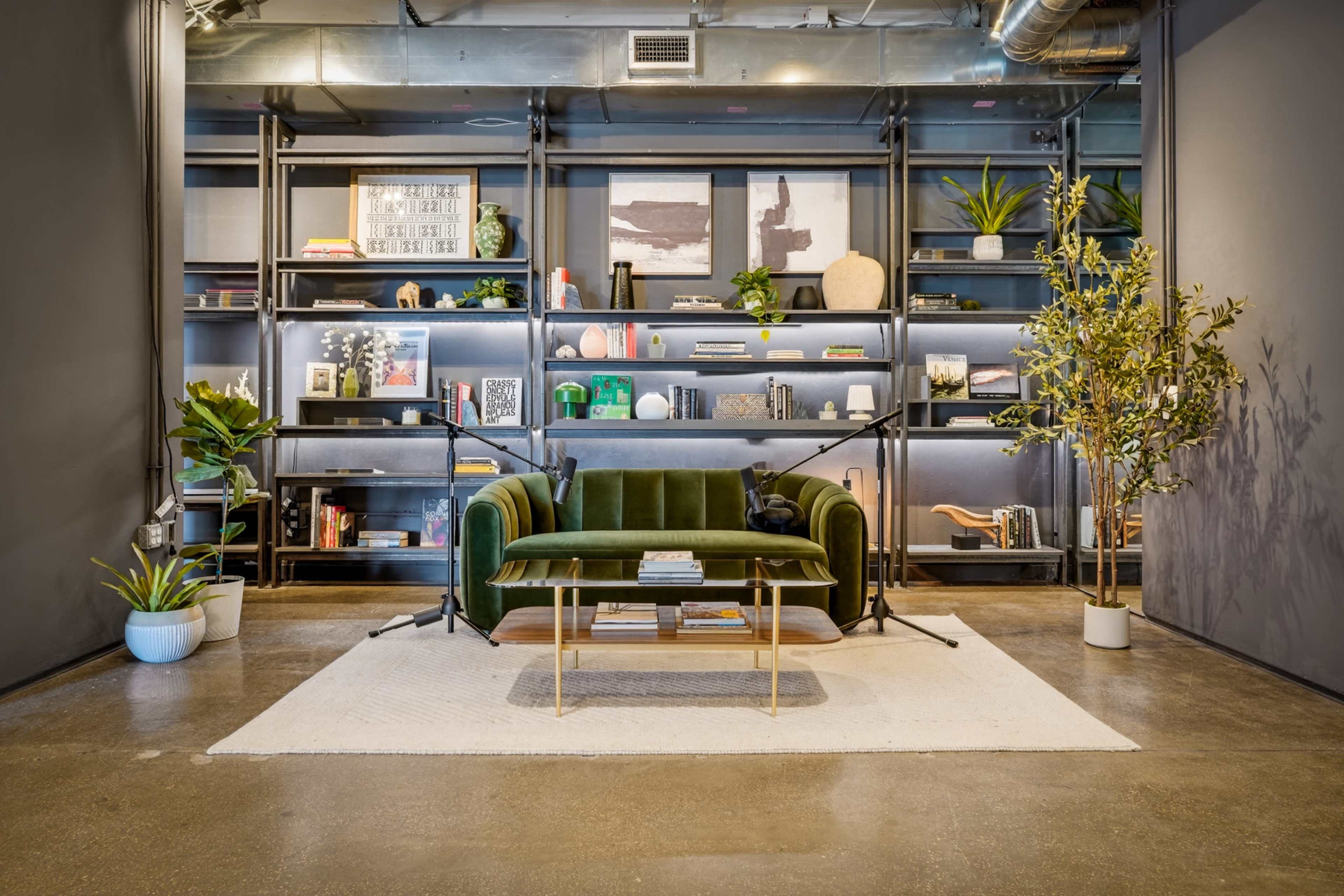 The image shows a modern living space with a green sofa facing a stylish coffee table, surrounded by shelves filled with books and decorative items, and adorned with plants.