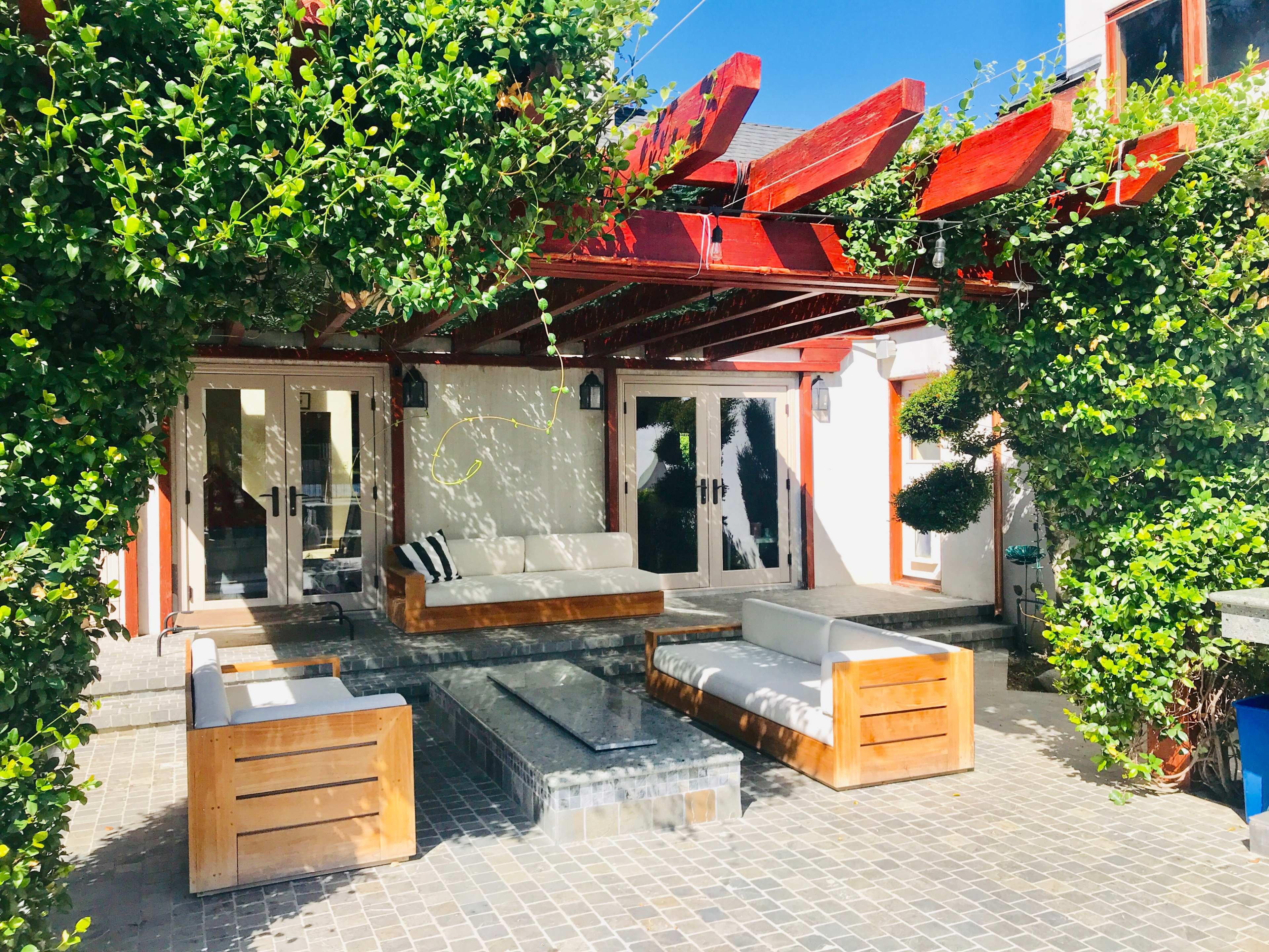 A patio features a seating area with white sofas and a table surrounded by greenery and wooden beams.