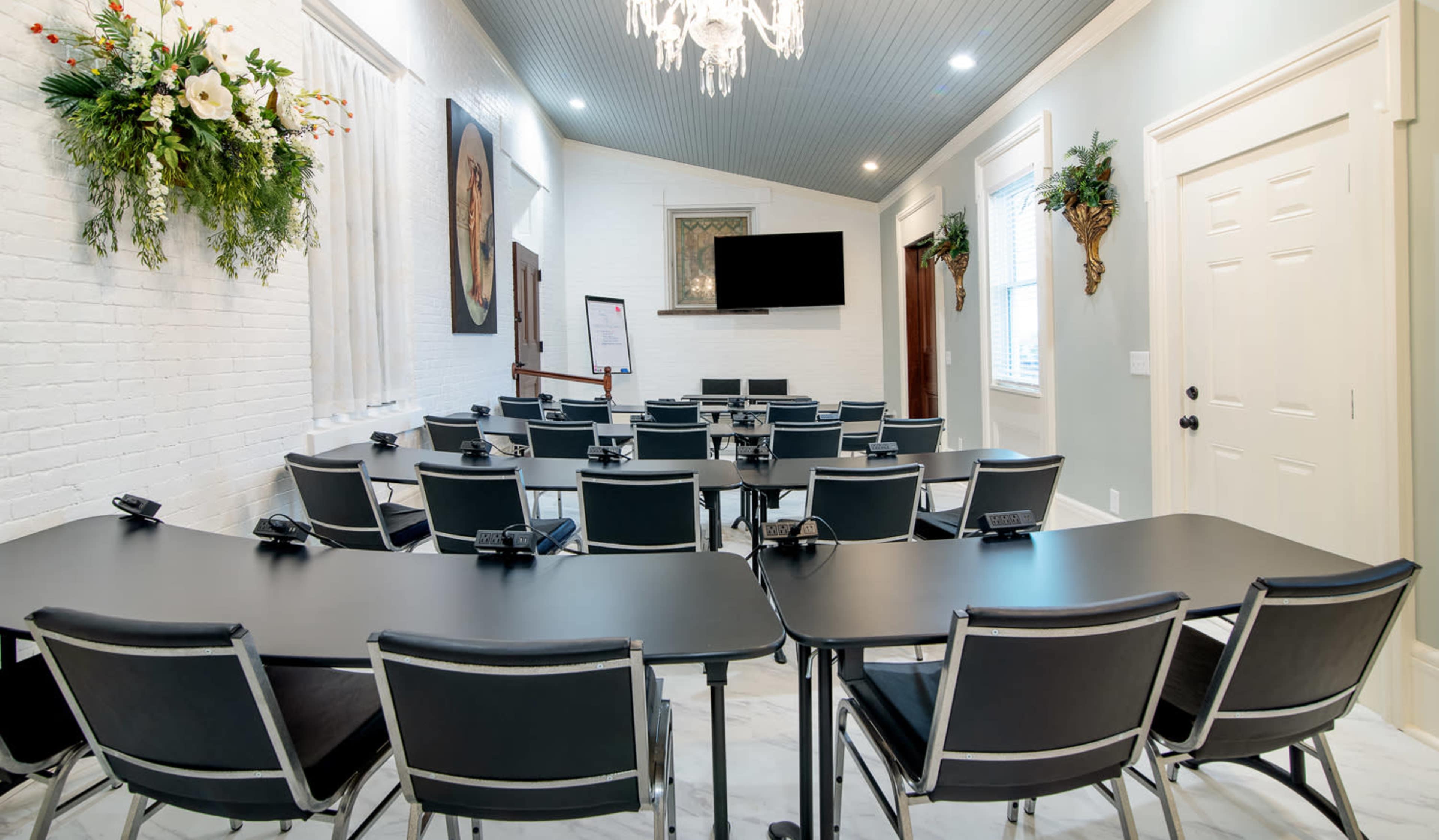 A well-lit conference room arranged with black tables and chairs, featuring a large screen and a whiteboard on one wall.