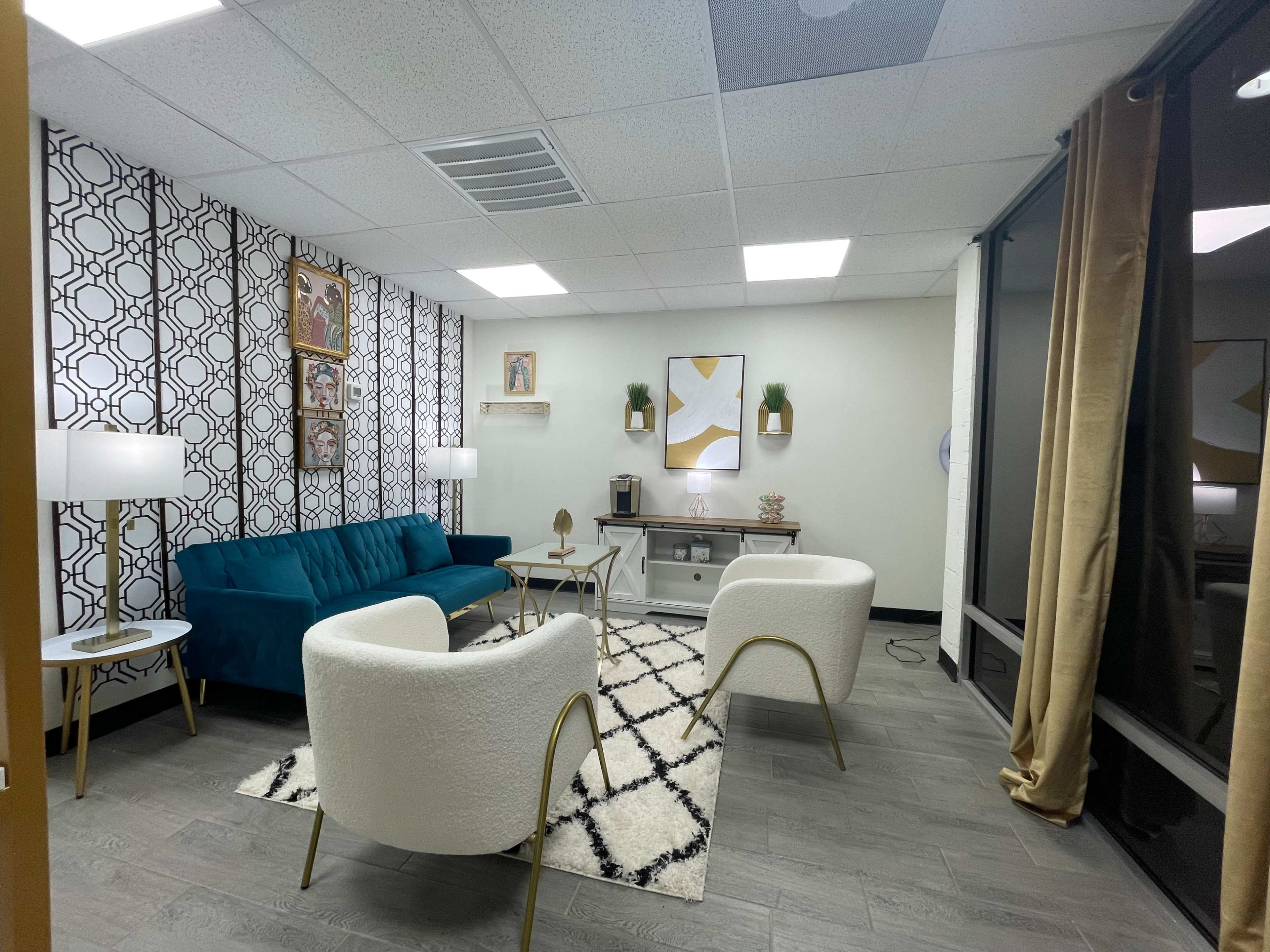 A modern lounge area featuring a teal sofa, two white chairs, a coffee table, and decorative wall art against a patterned backdrop.