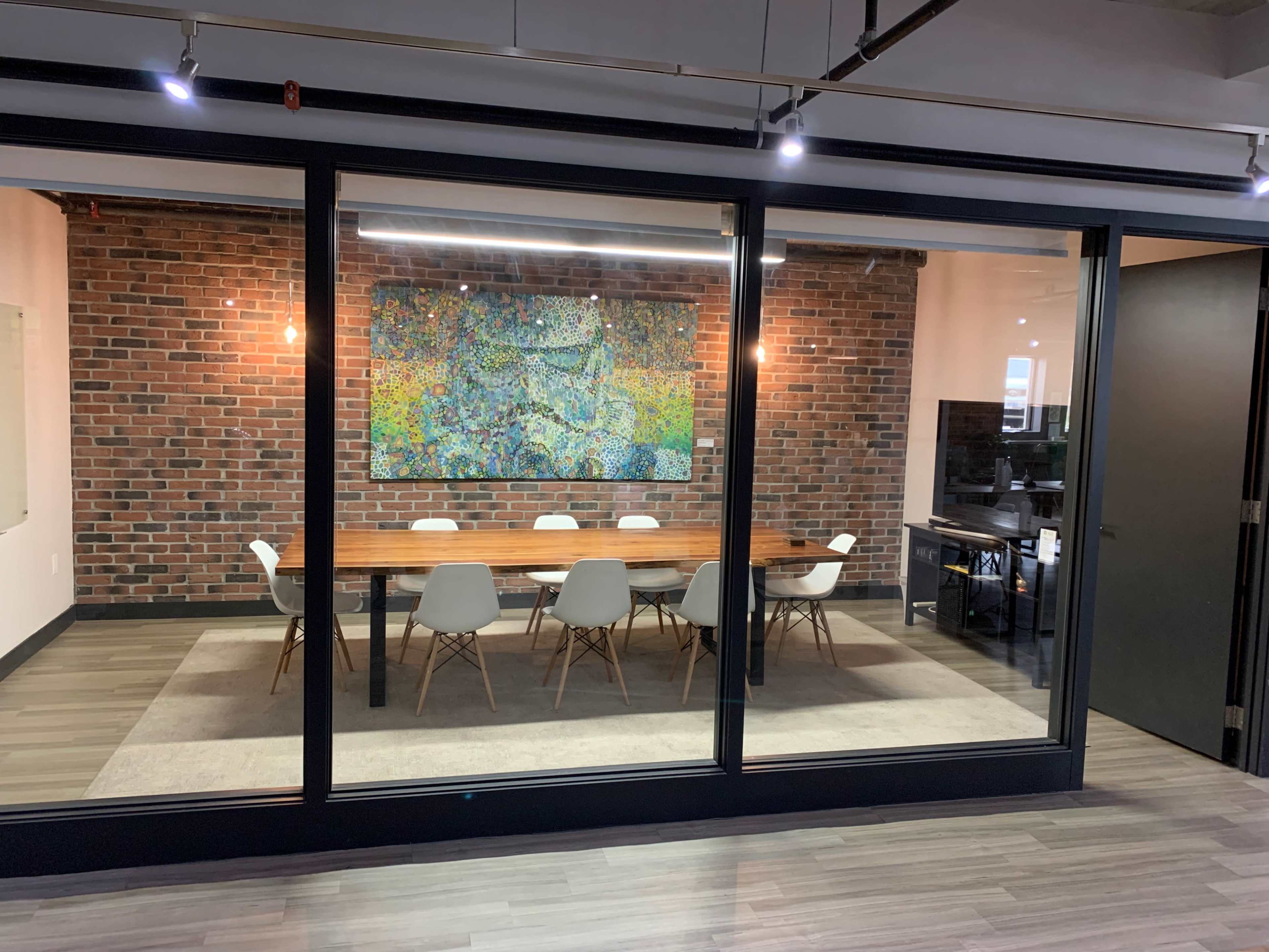 A conference room features a large wooden table surrounded by white chairs, with a colorful abstract painting on the brick wall.