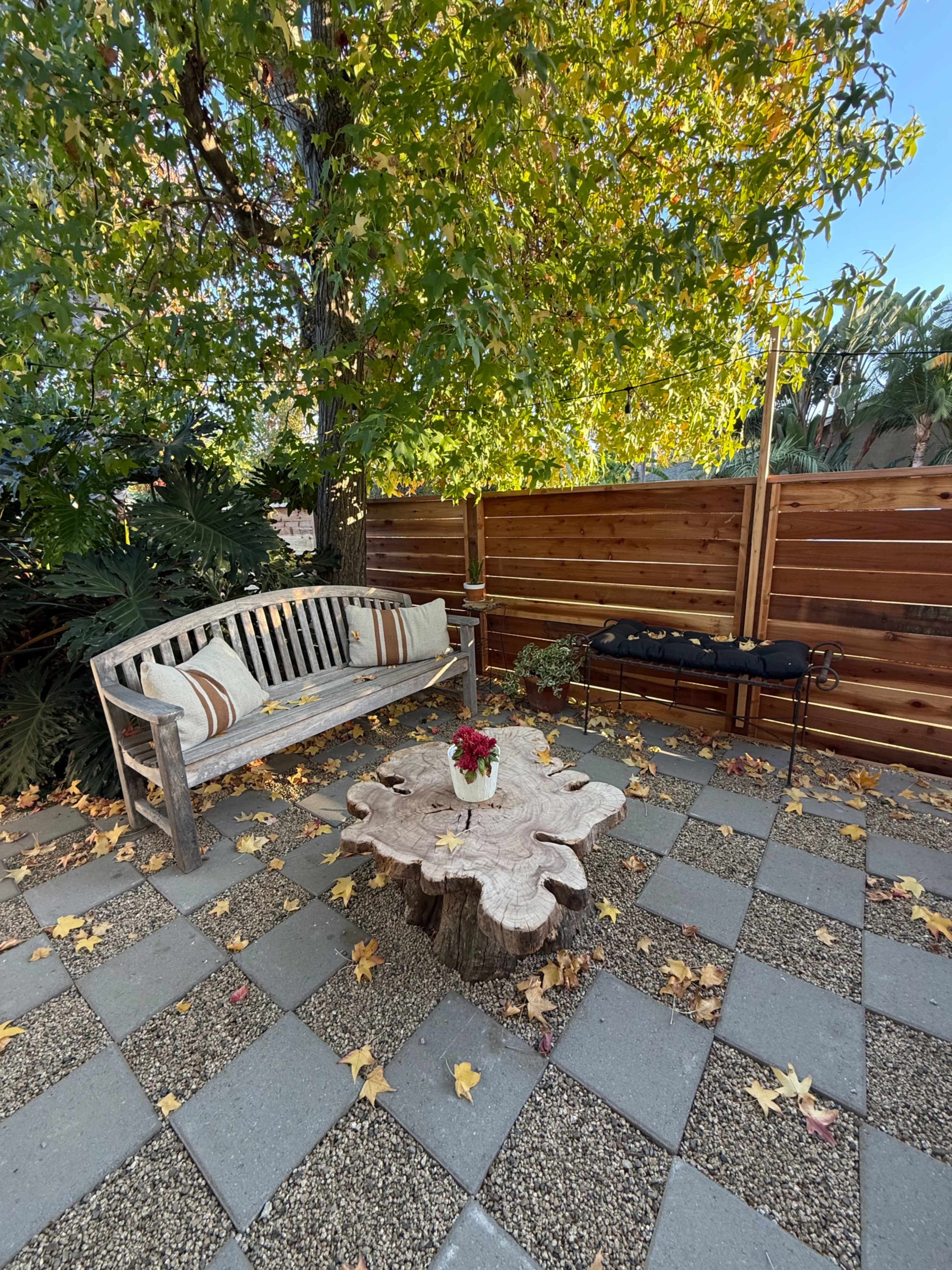 A wooden bench sits beside a round, tree stump table on a patio covered with pavers, surrounded by autumn leaves and a wooden fence.