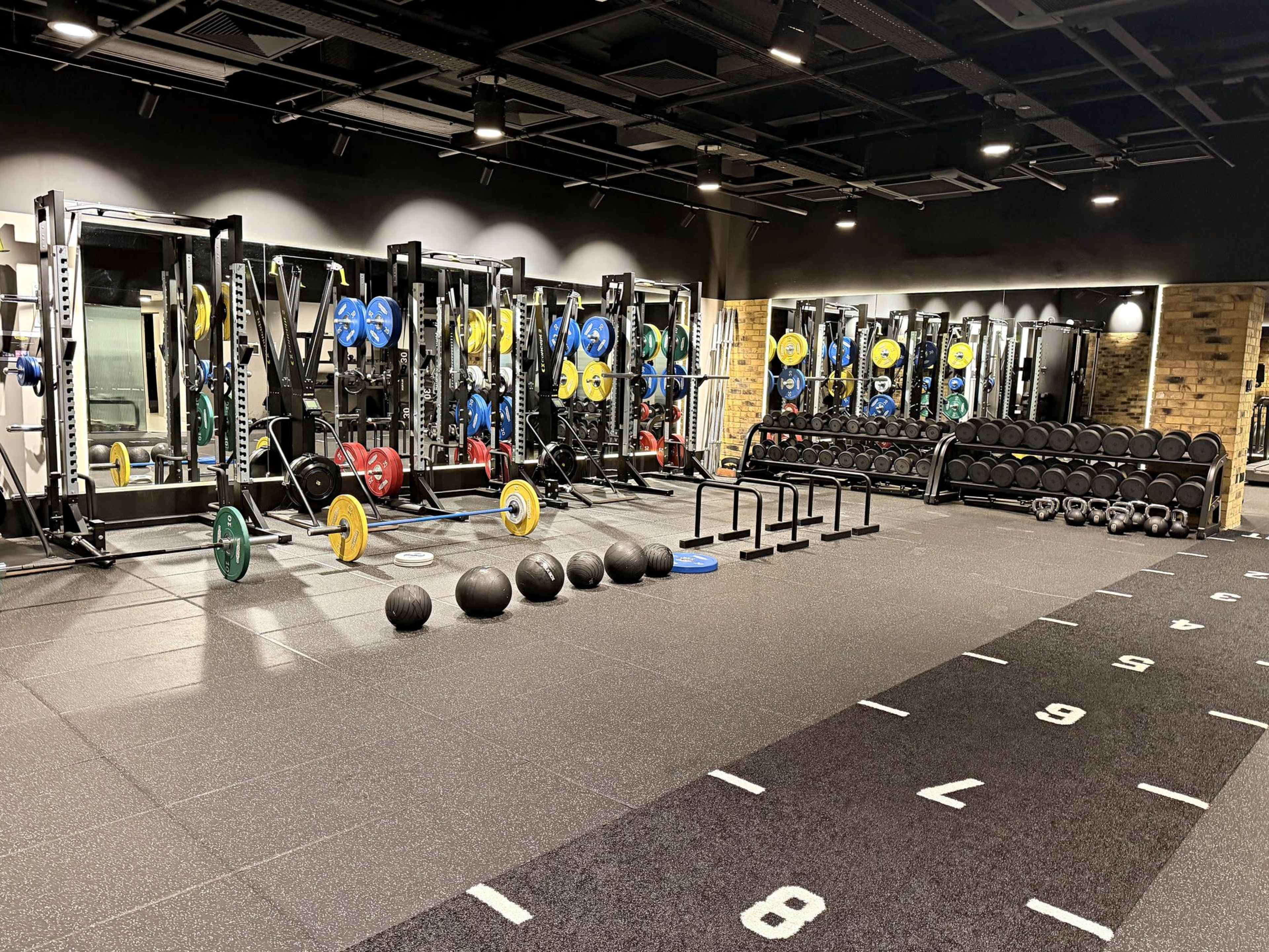 The image shows a well-equipped gym featuring weightlifting equipment, including racks, barbells, dumbbells, and various fitness accessories.