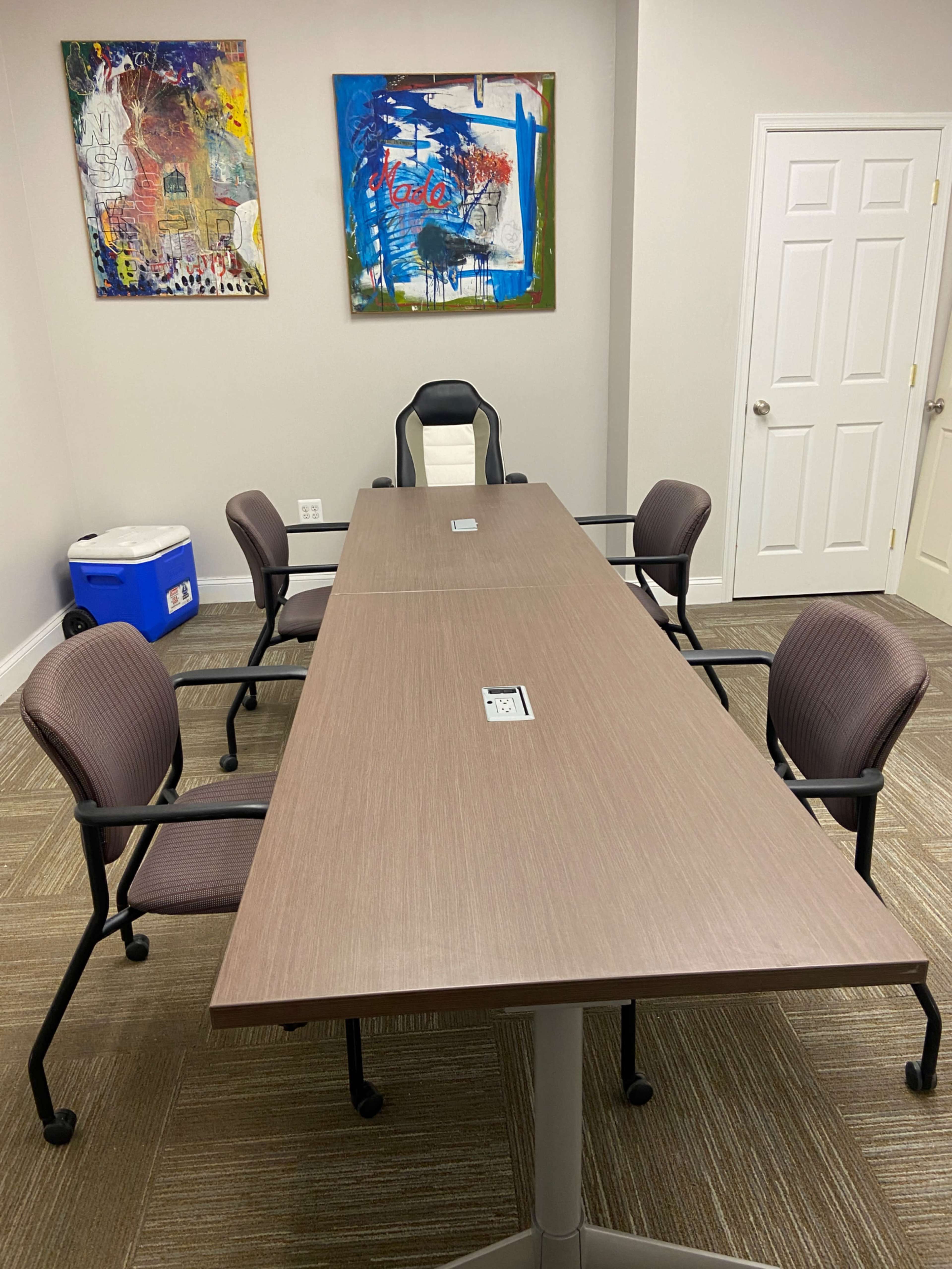 A modern conference room with a large table surrounded by four chairs, and two abstract paintings on the wall.