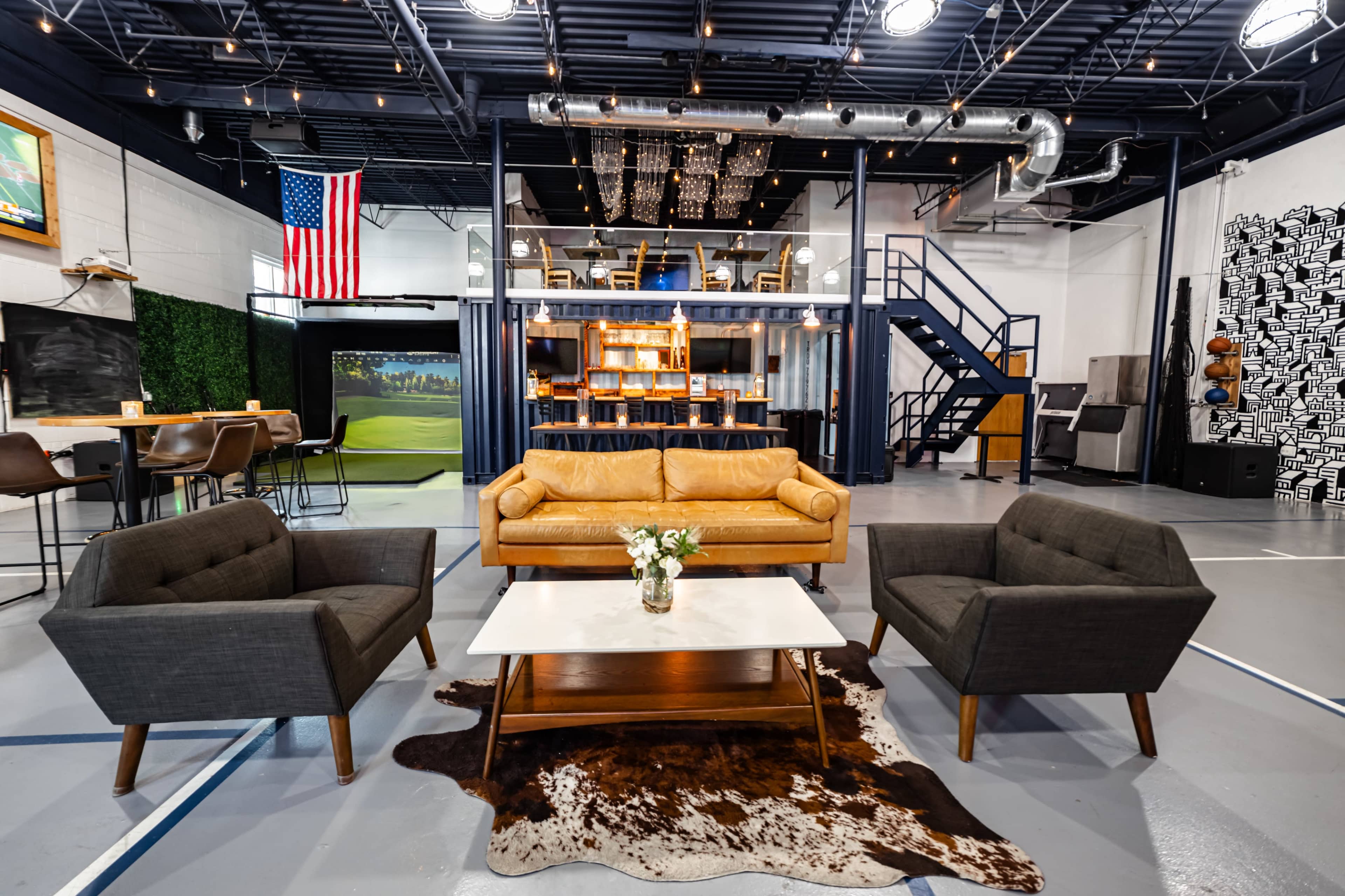 A spacious modern lounge area with a yellow leather couch, two gray armchairs, a coffee table on a cowhide rug, and a staircase leading to an upper level.