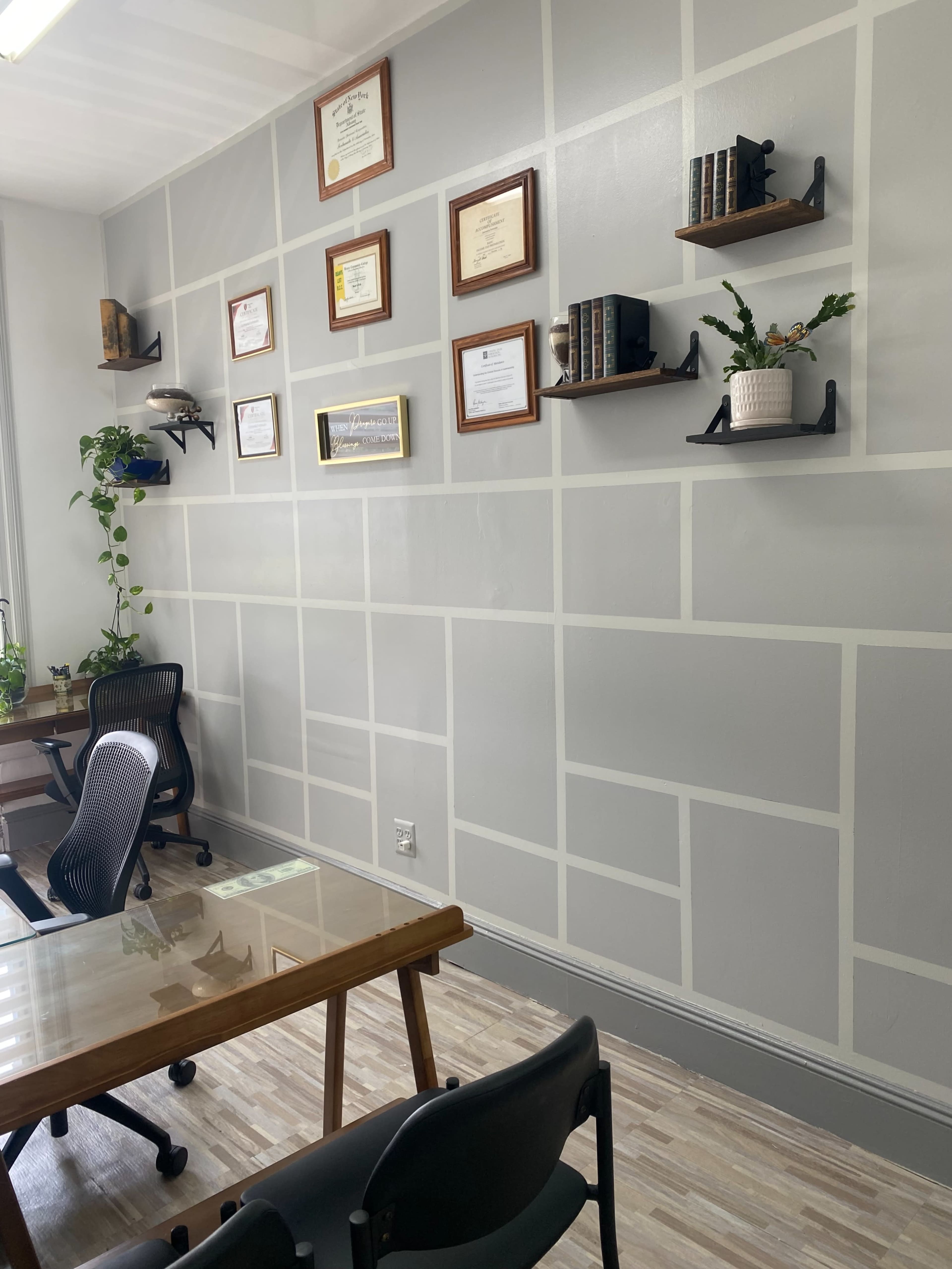 A modern office space with a wall featuring framed certificates and books on shelves, alongside a glass table and black chairs.