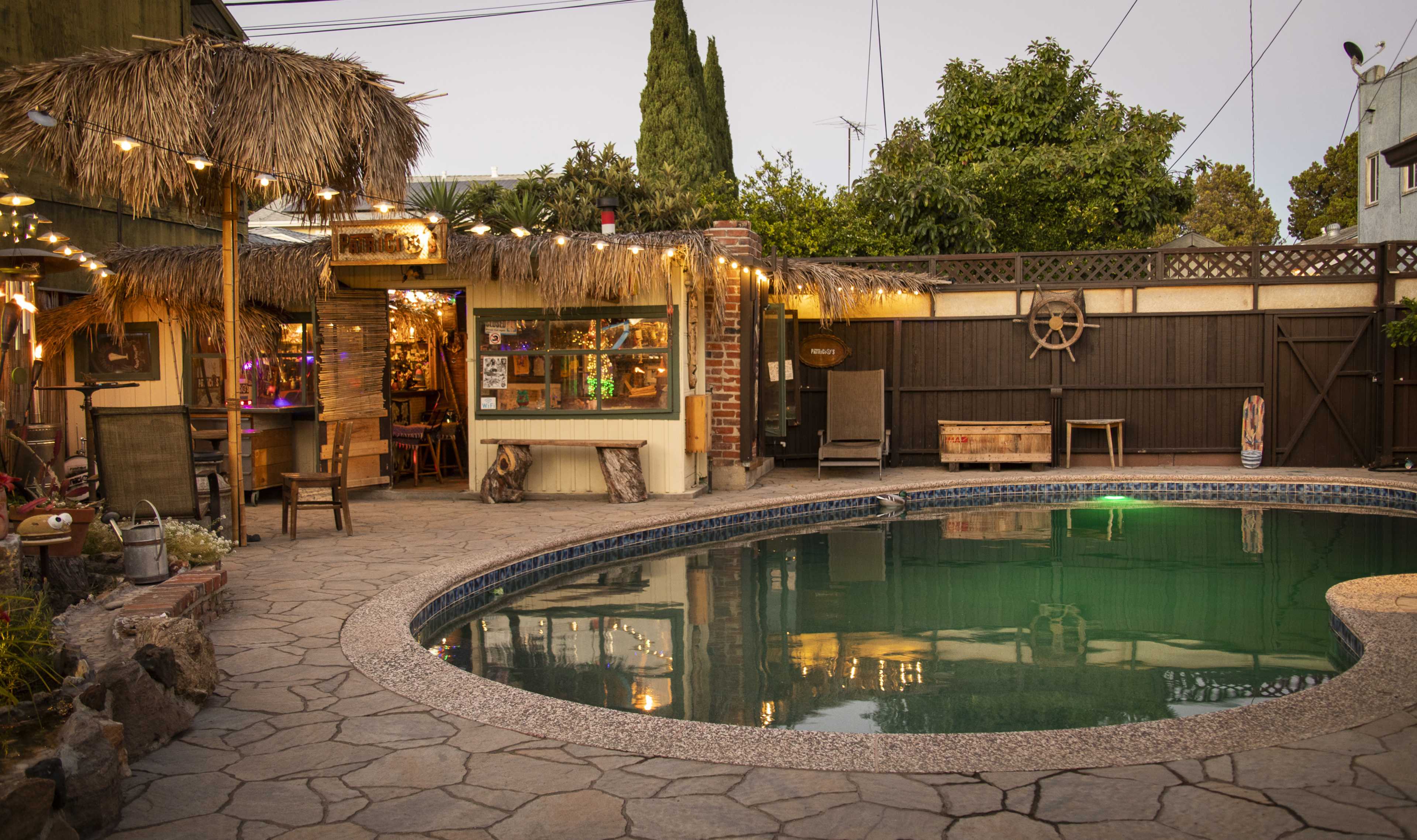 The scene features a backyard with a pool surrounded by stone paving, a thatched-roof bar area, and string lights overhead.
