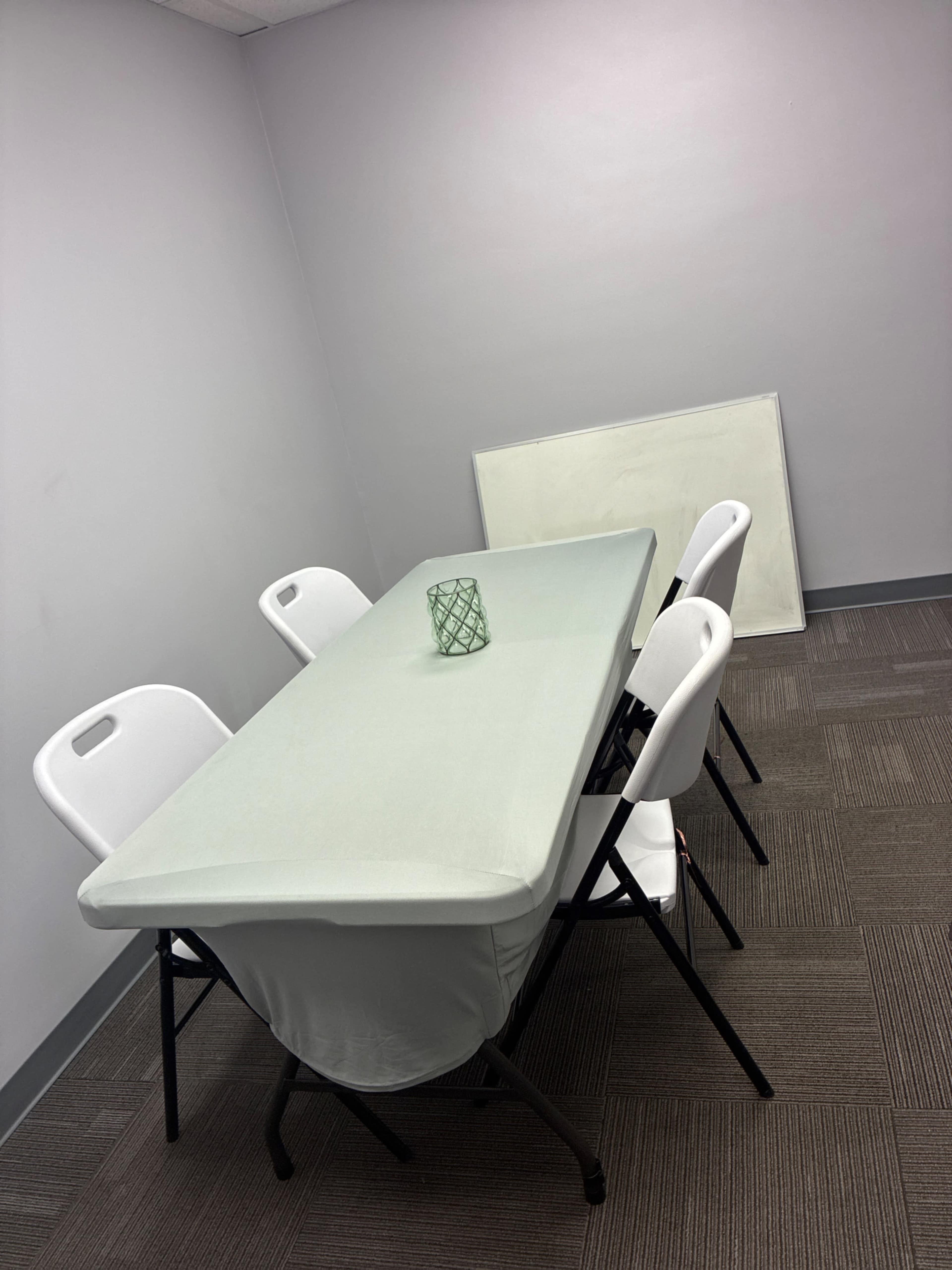 A light green tablecloth covers a rectangular folding table surrounded by four white plastic chairs in a sparsely furnished room.