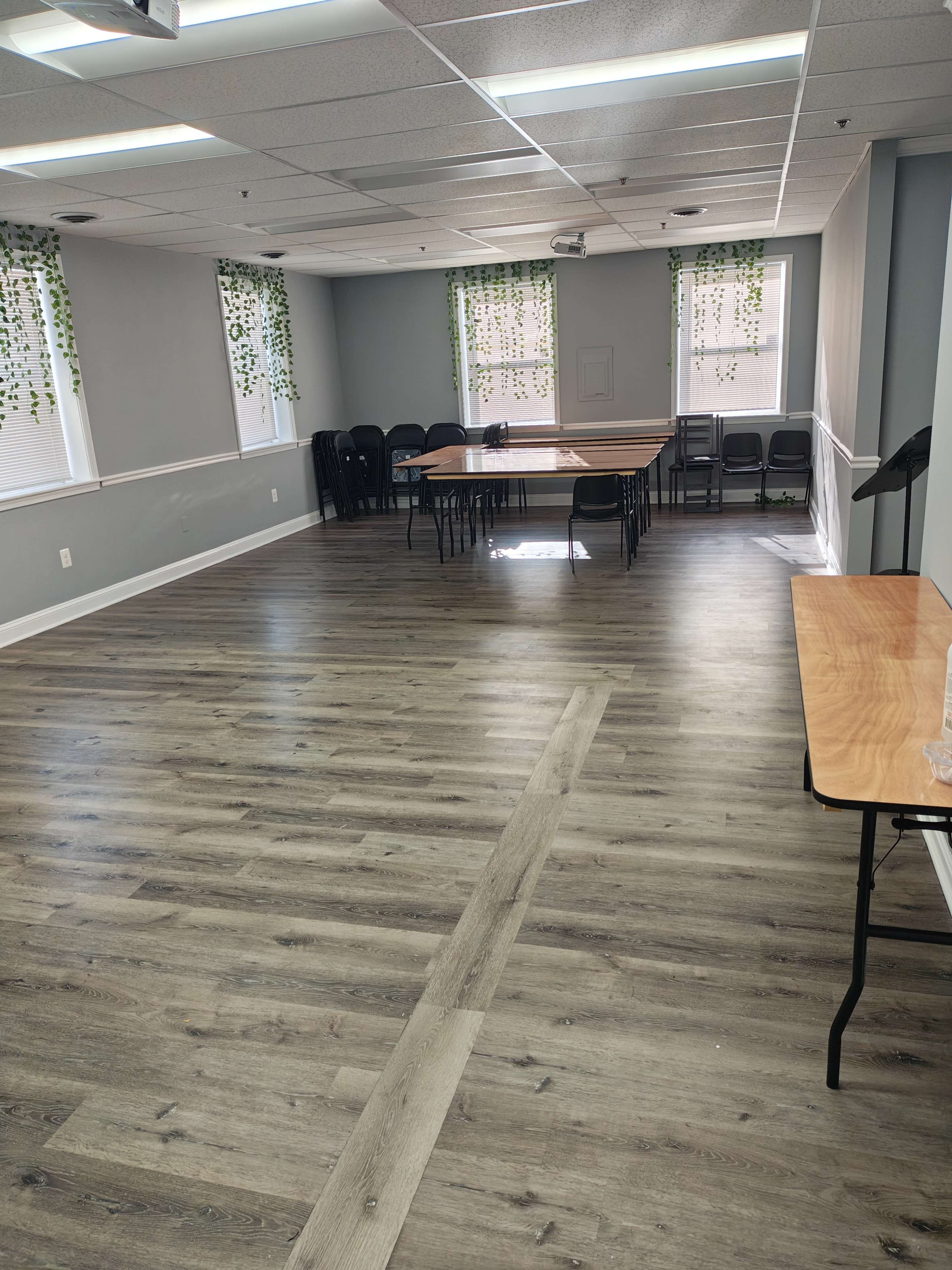 The image shows a spacious meeting room with wooden flooring, a large rectangular table surrounded by black chairs, and green vines hanging from the windows.
