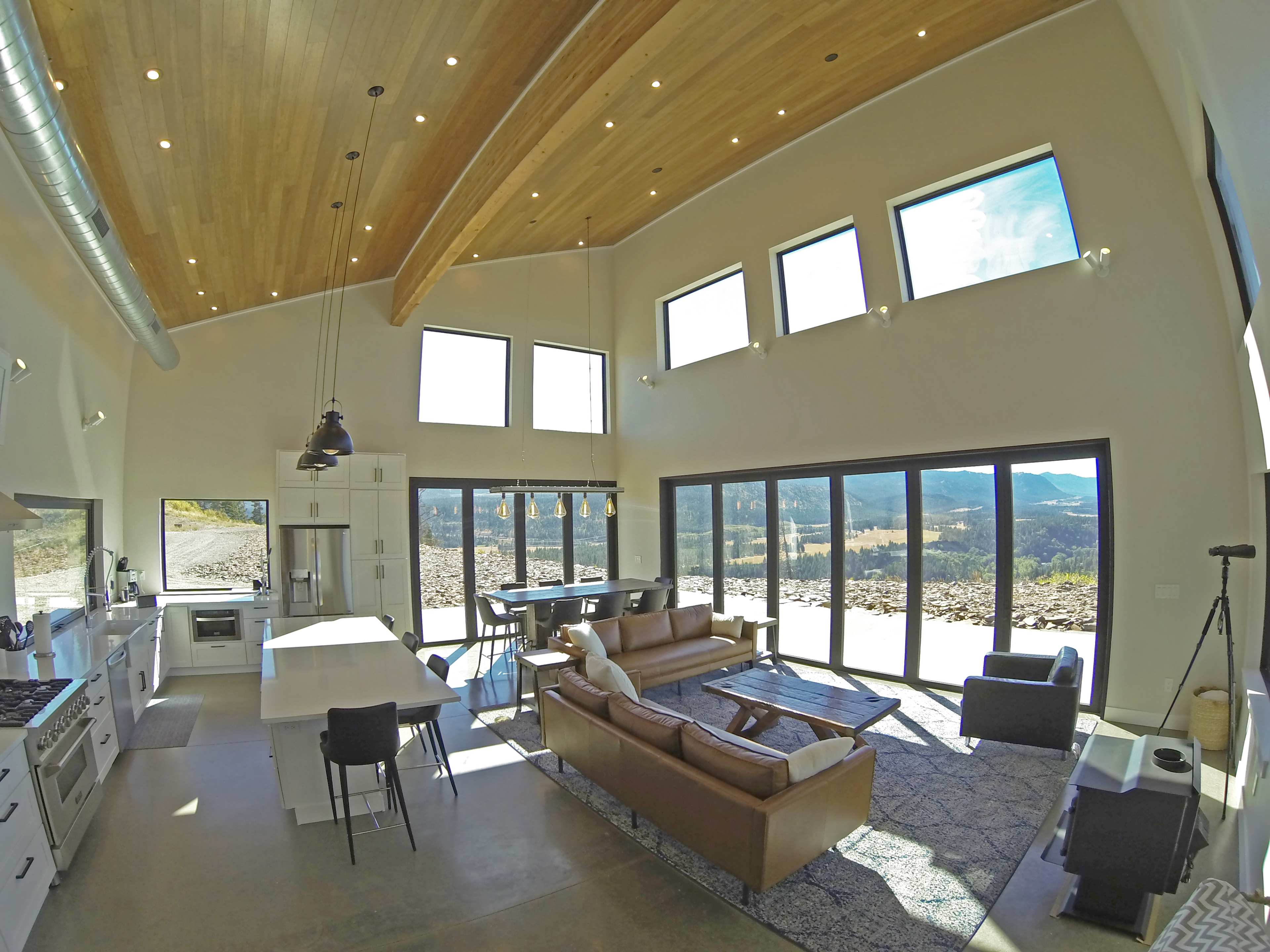 A modern, open-concept living space with a kitchen, dining area, and seating area, featuring large windows that provide a view of the surrounding landscape.