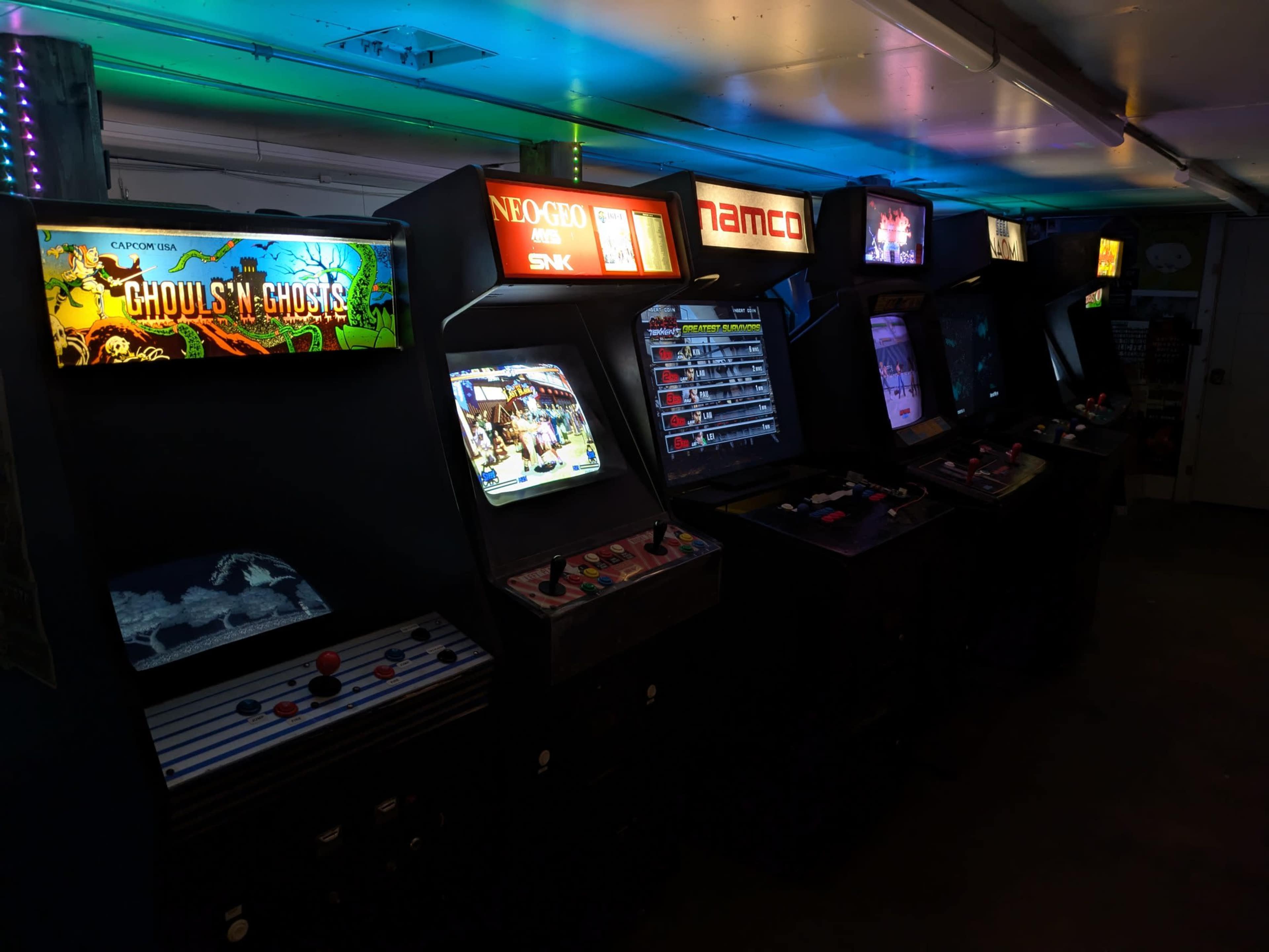 The image shows a row of vintage arcade game machines illuminated by colorful lights, with various game titles displayed on their screens.