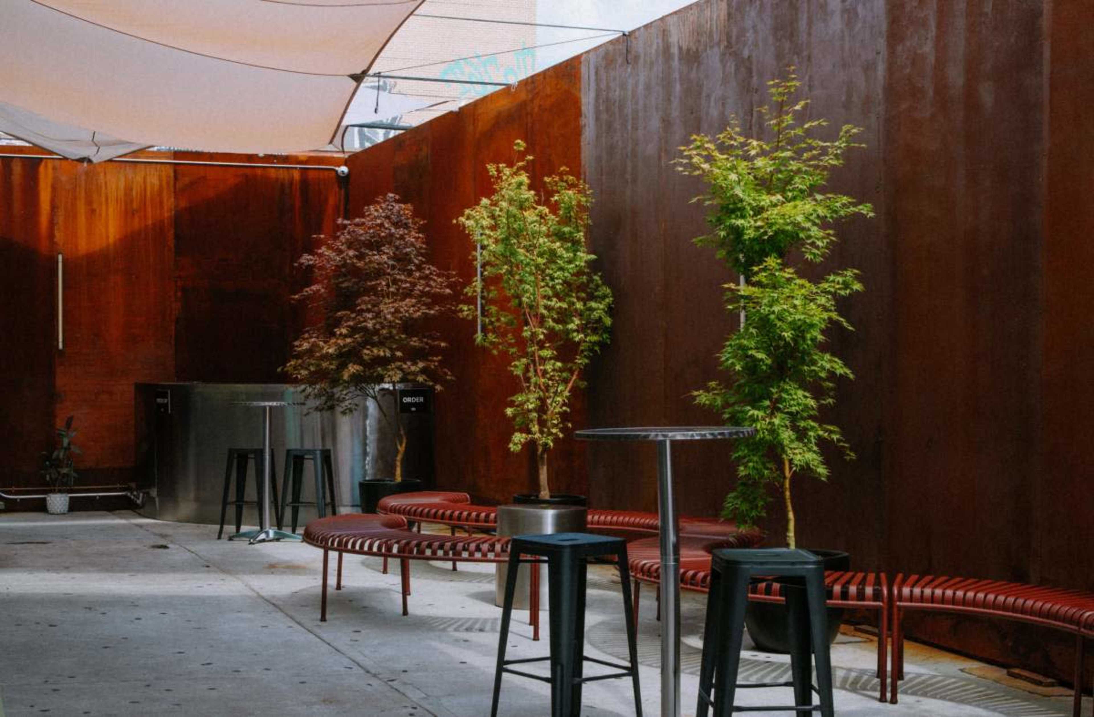 The image shows a modern outdoor space with metal walls, seating along the side, and three potted trees.