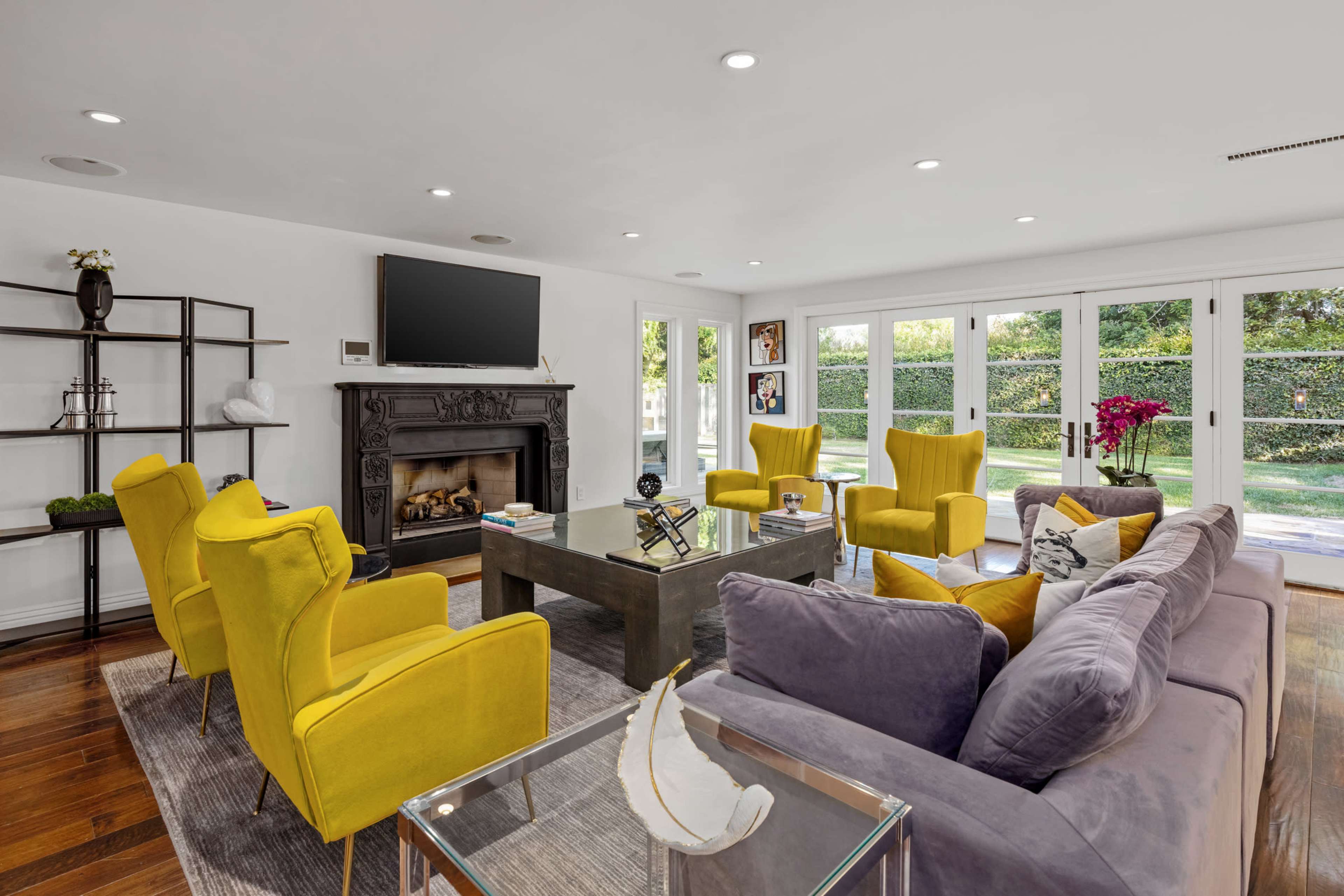 A modern living room features a gray sofa, yellow chairs, a coffee table, and a fireplace, with large windows that open to a green backyard.