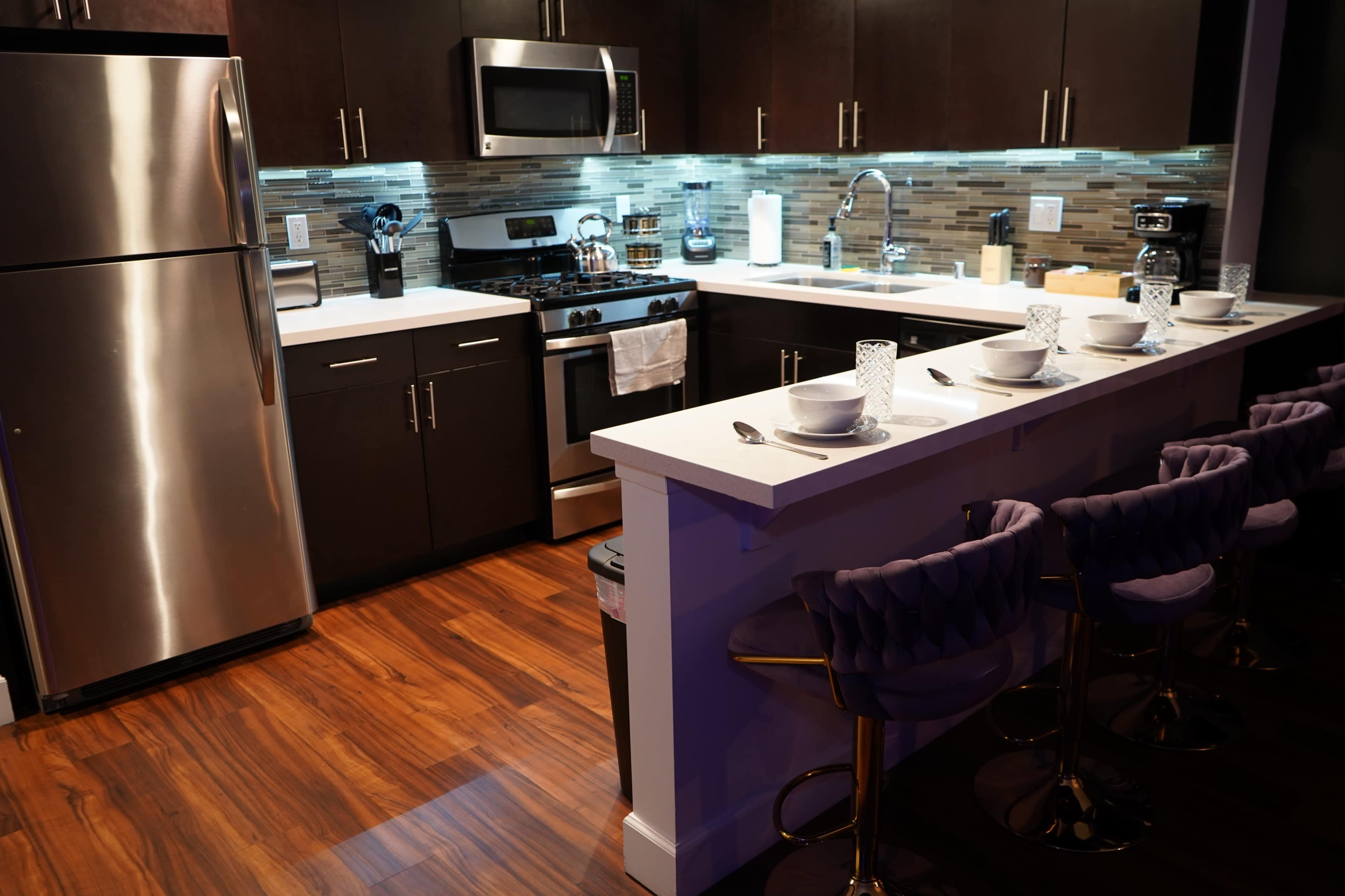 A modern kitchen features stainless steel appliances, dark cabinetry, a white countertop, and bar stools arranged at an island.