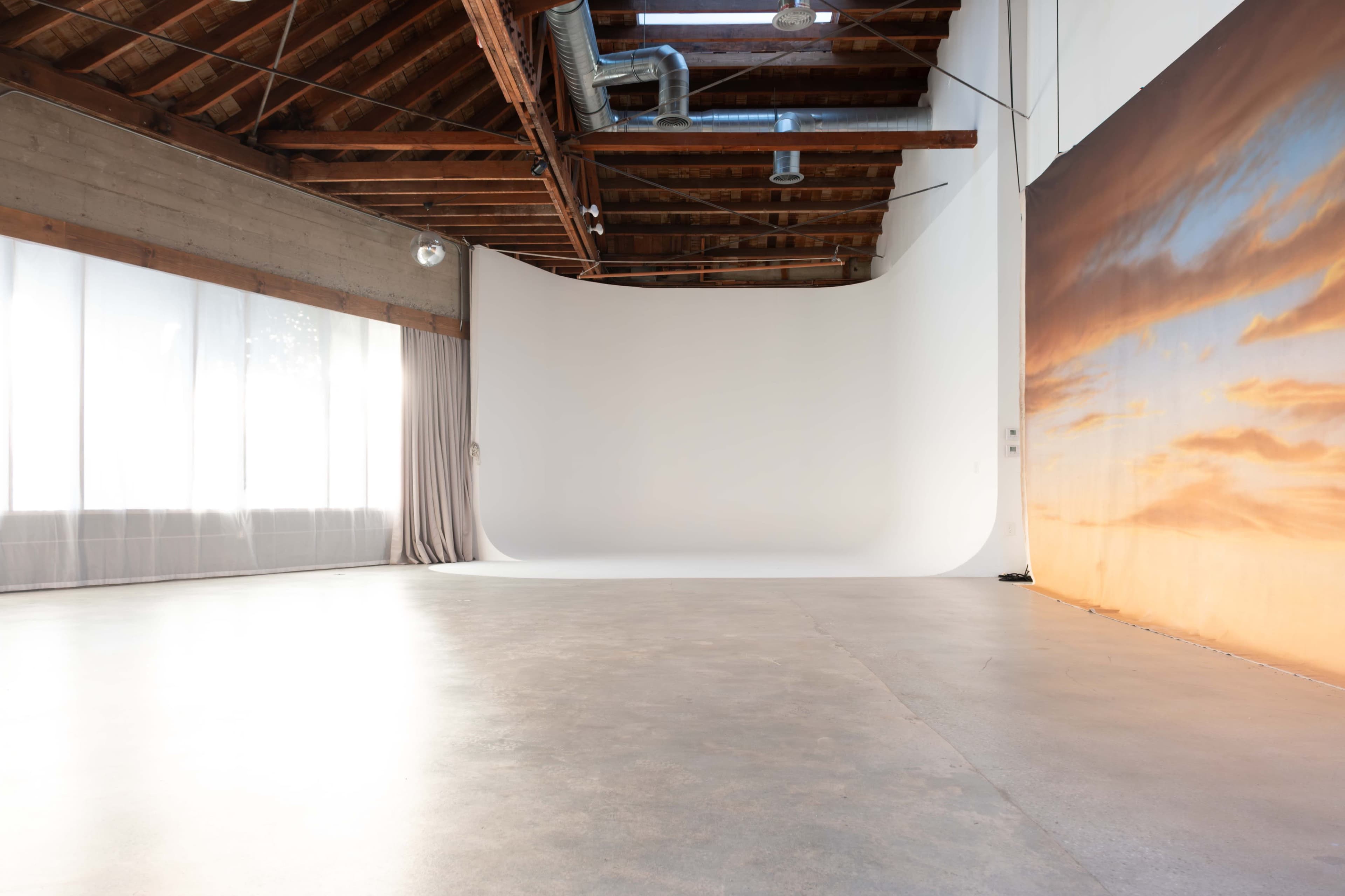 The image shows a spacious photography studio with a white backdrop, concrete flooring, and wooden beams overhead.