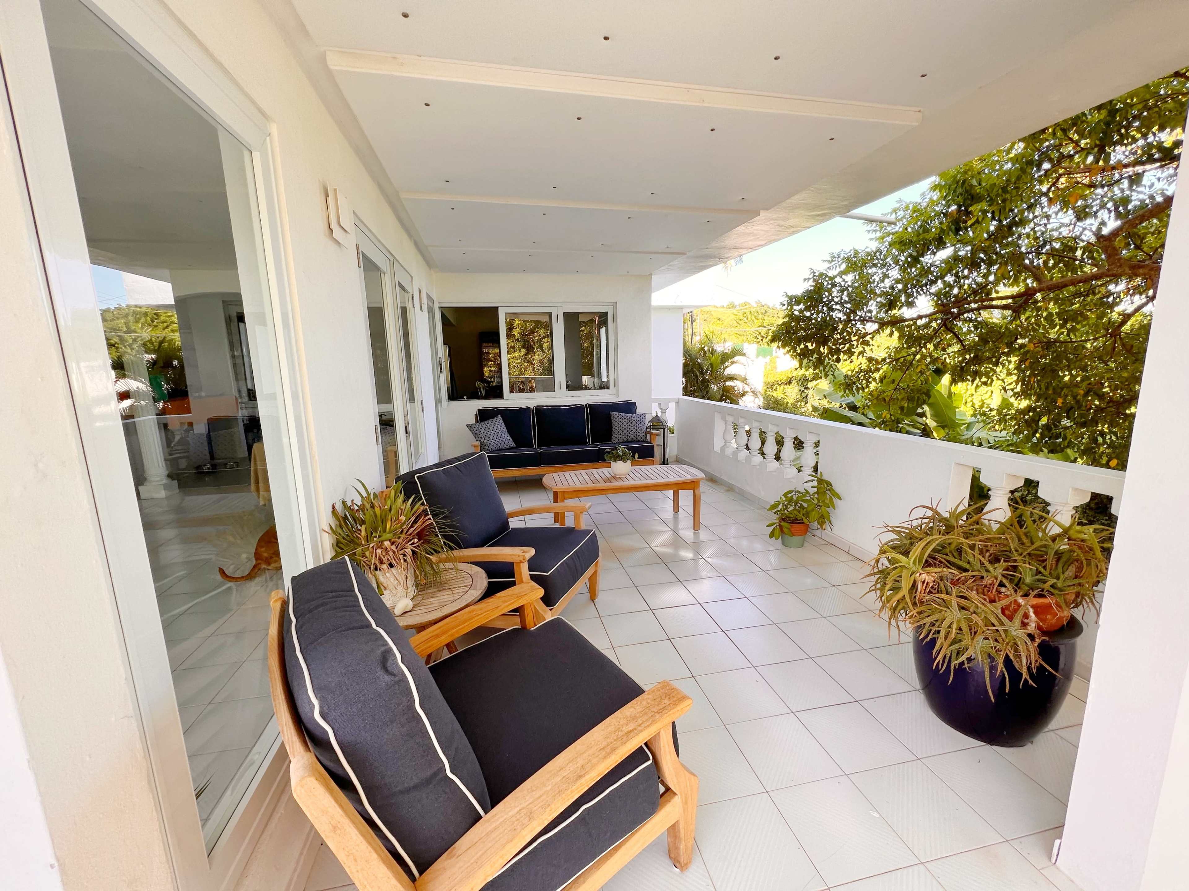 The image depicts a balcony with wooden and cushioned seating arrangements, a coffee table, and potted plants, surrounded by white walls and large glass doors.