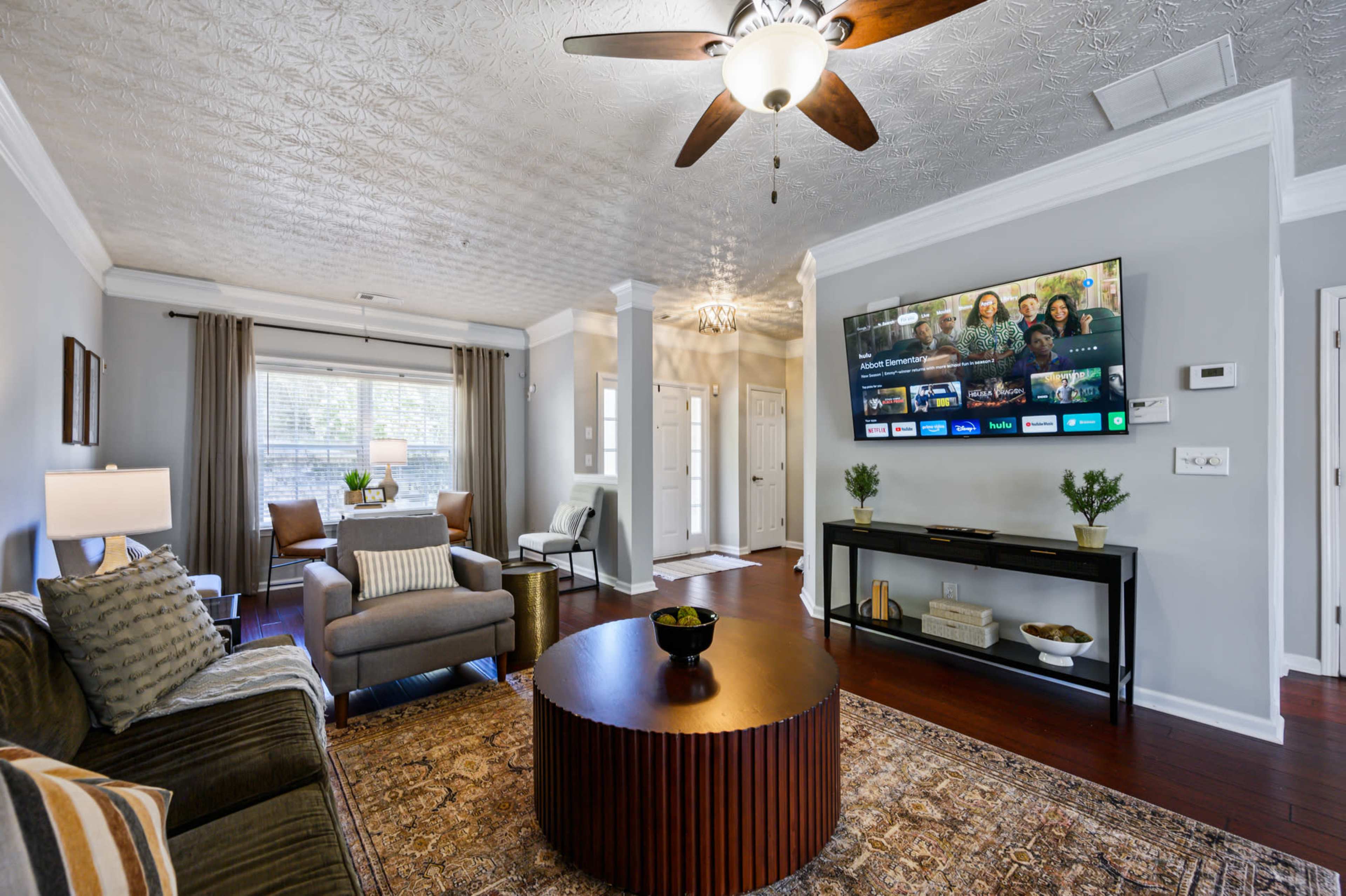 The living room features a gray sofa, a round wooden coffee table, and a large television mounted on the wall, with a window allowing natural light.