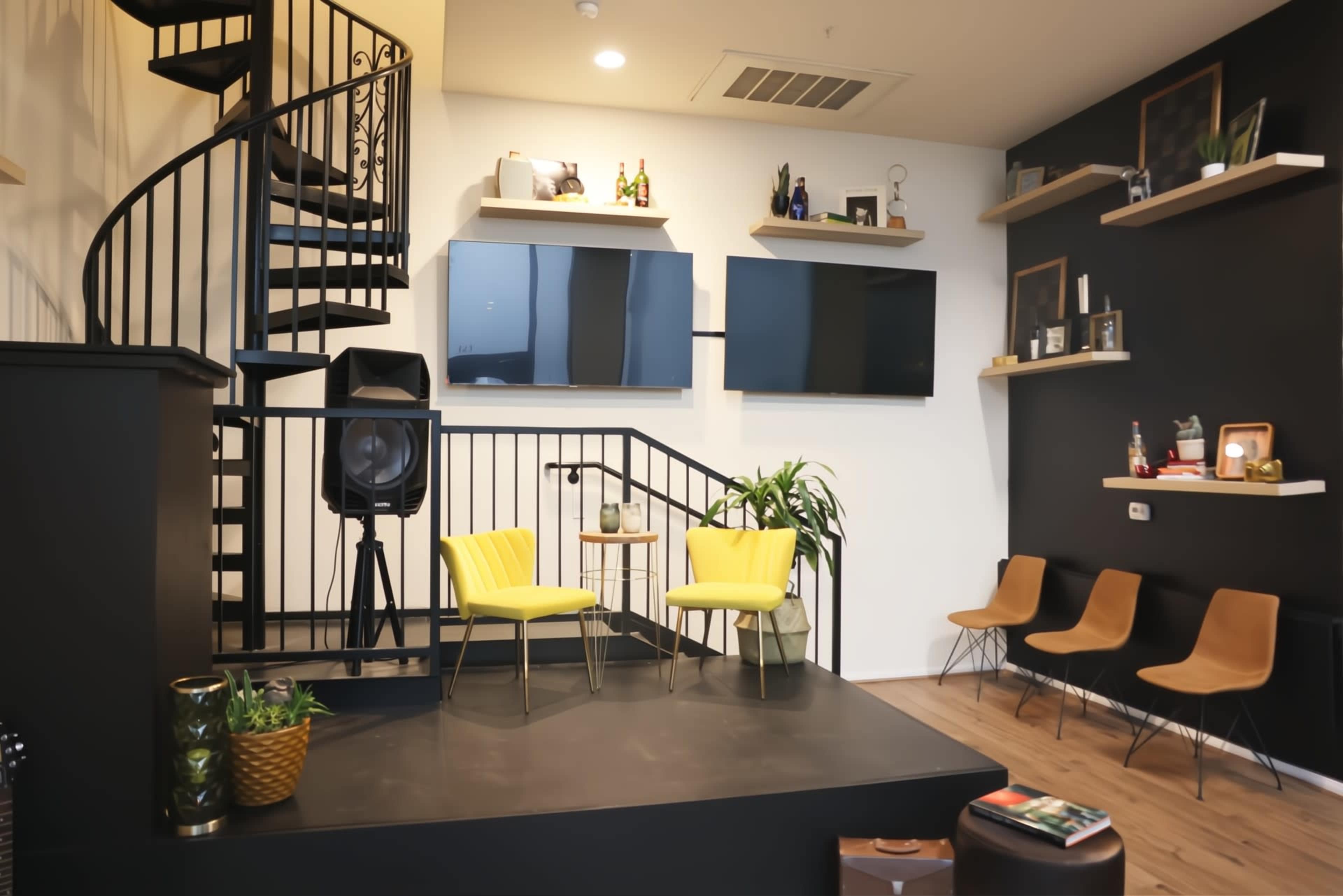 A spiral staircase leads to an upper level in a modern room featuring two yellow chairs on a raised platform, with audio equipment and decorative shelves on the walls.