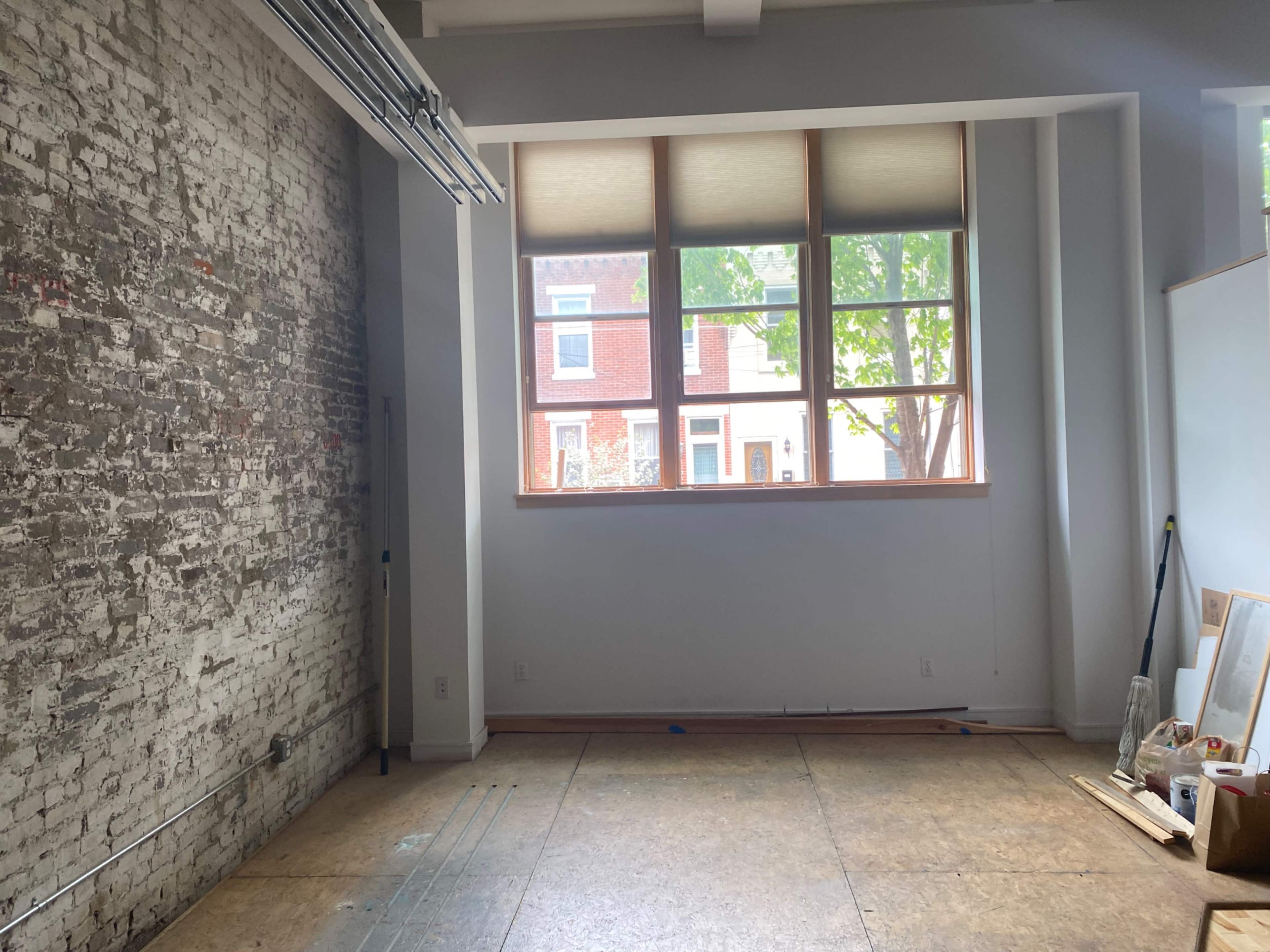 The image shows an empty room with a brick wall on one side and large windows overlooking a street with houses outside.
