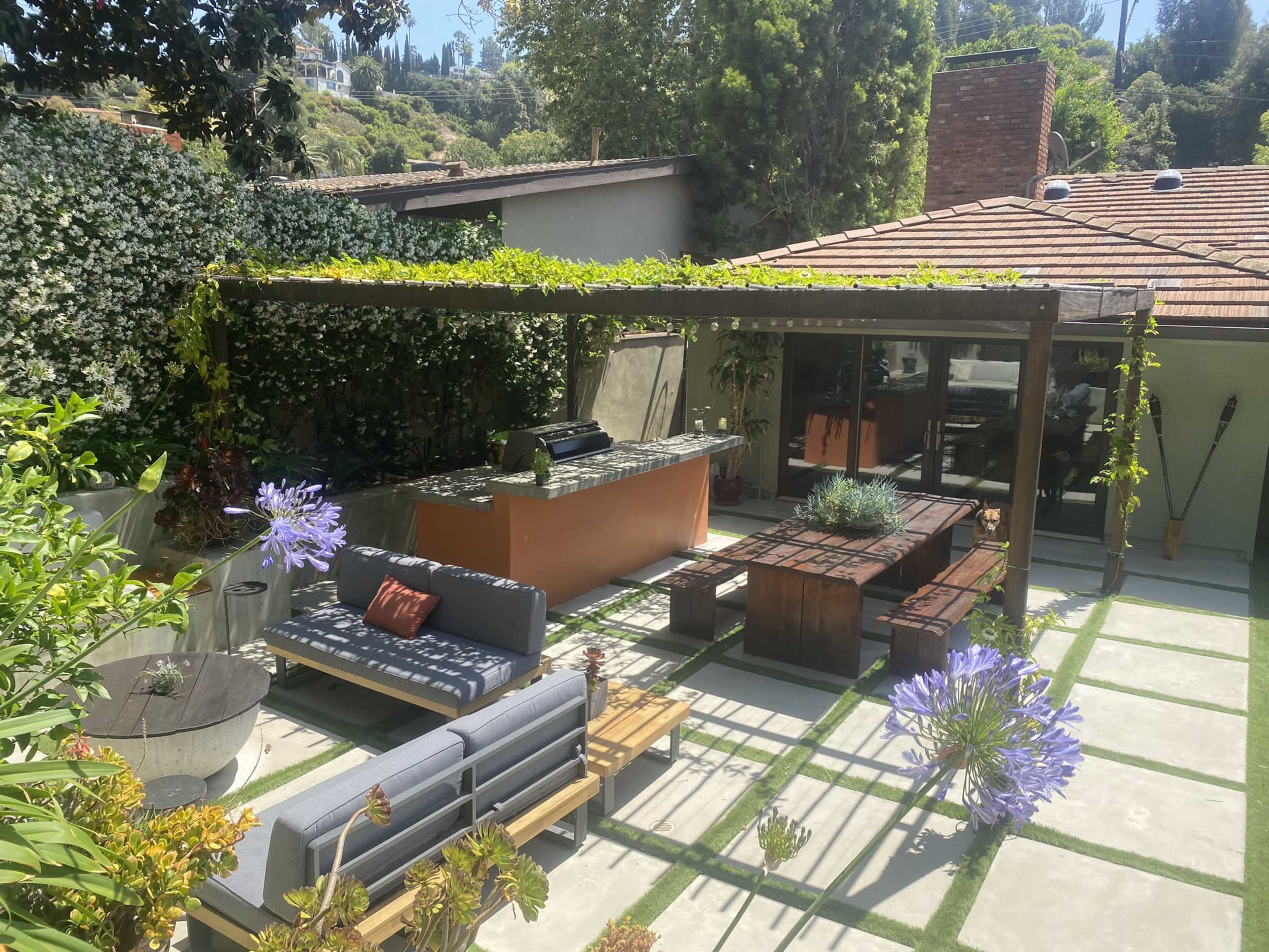 A backyard patio with a dining area, outdoor kitchen, and seating arrangements surrounded by greenery and flowering plants.