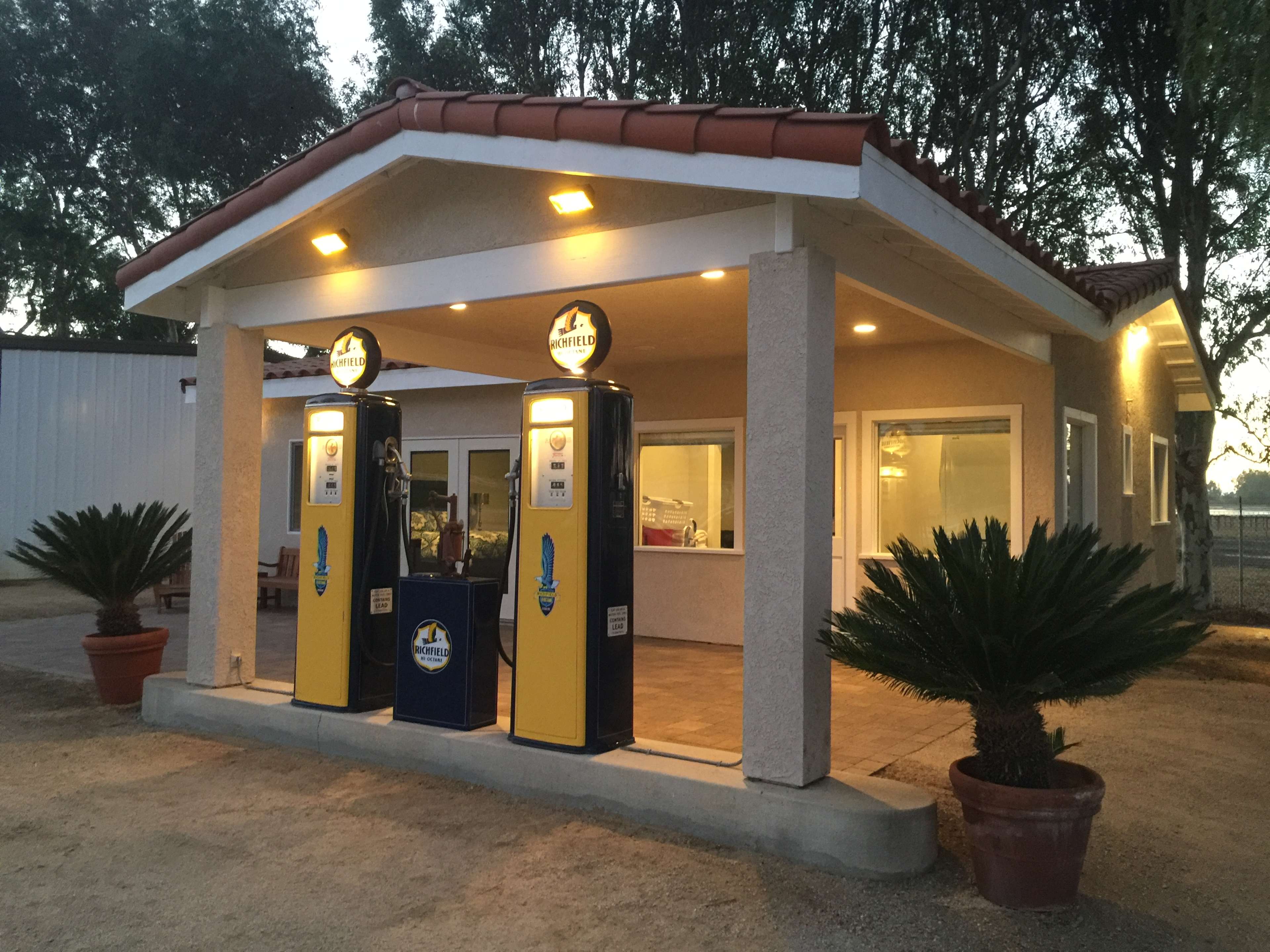 A small gas station features two vintage gas pumps in front of a building with a tiled roof and large windows.