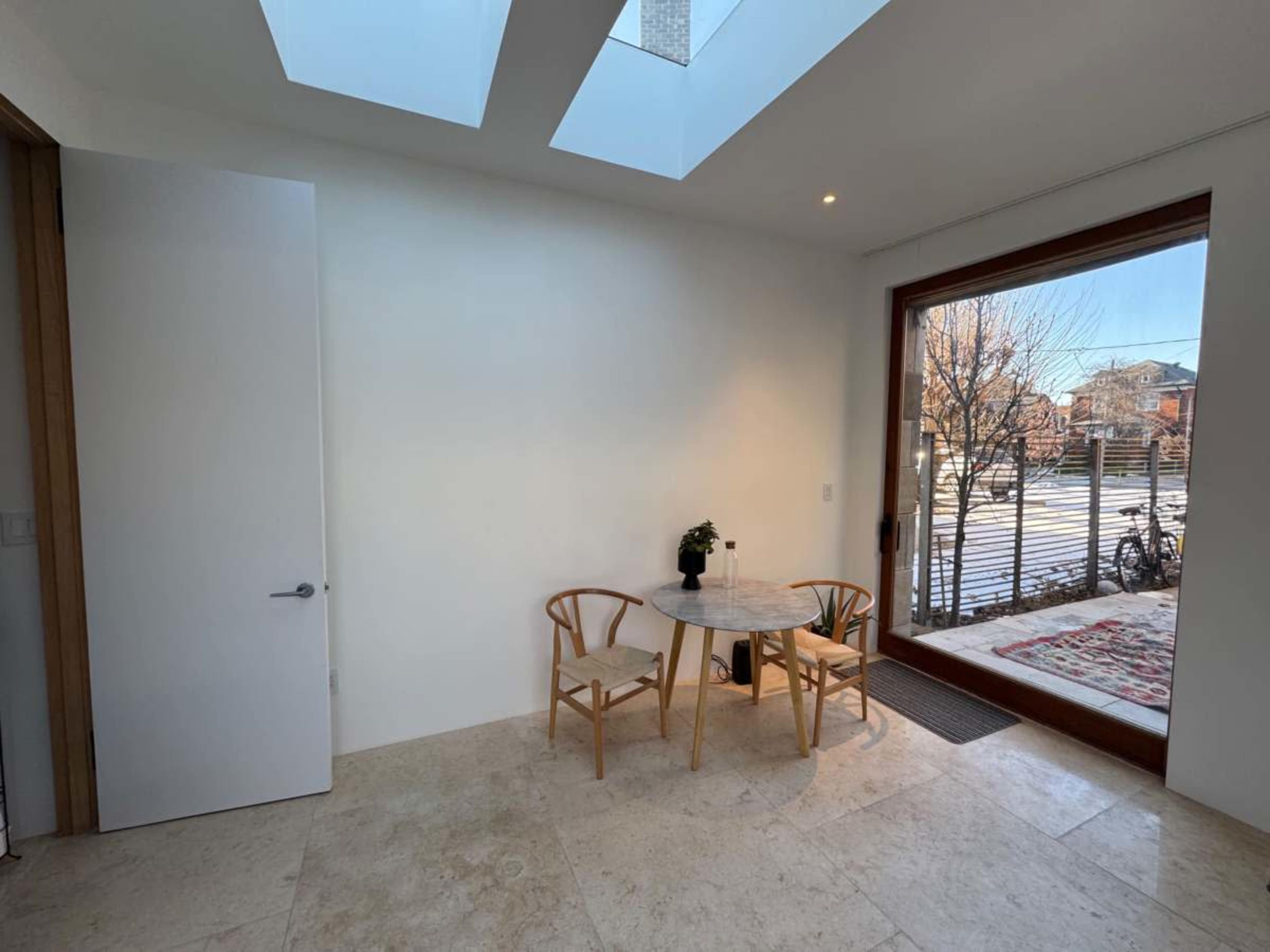 The image shows a minimalist indoor space featuring a small round table with two chairs, a potted plant, and large windows that provide natural light and a view of an outdoor area.