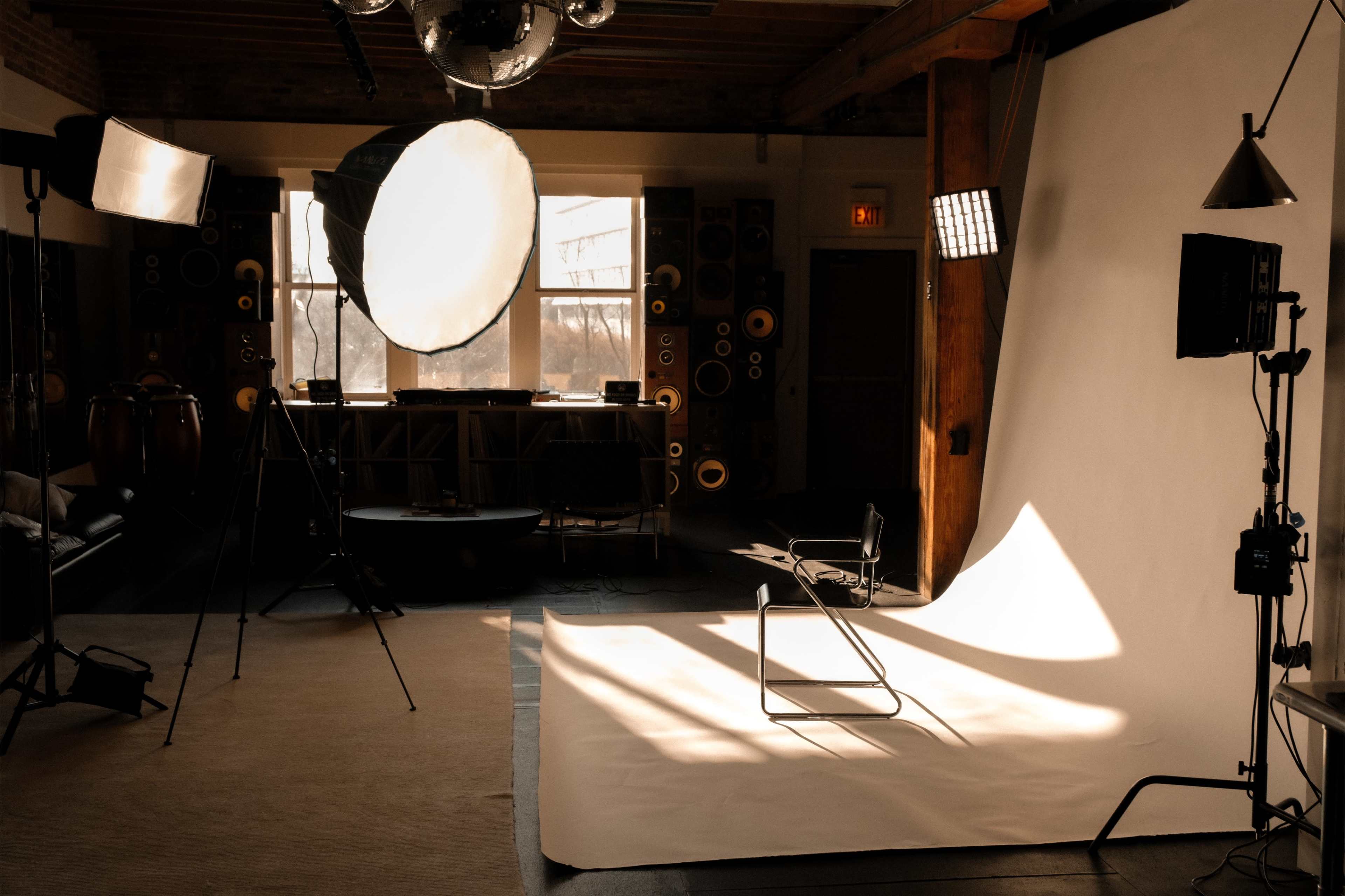 A photography studio features a plain backdrop, a chair, various lighting equipment, and sound equipment arranged in the background.