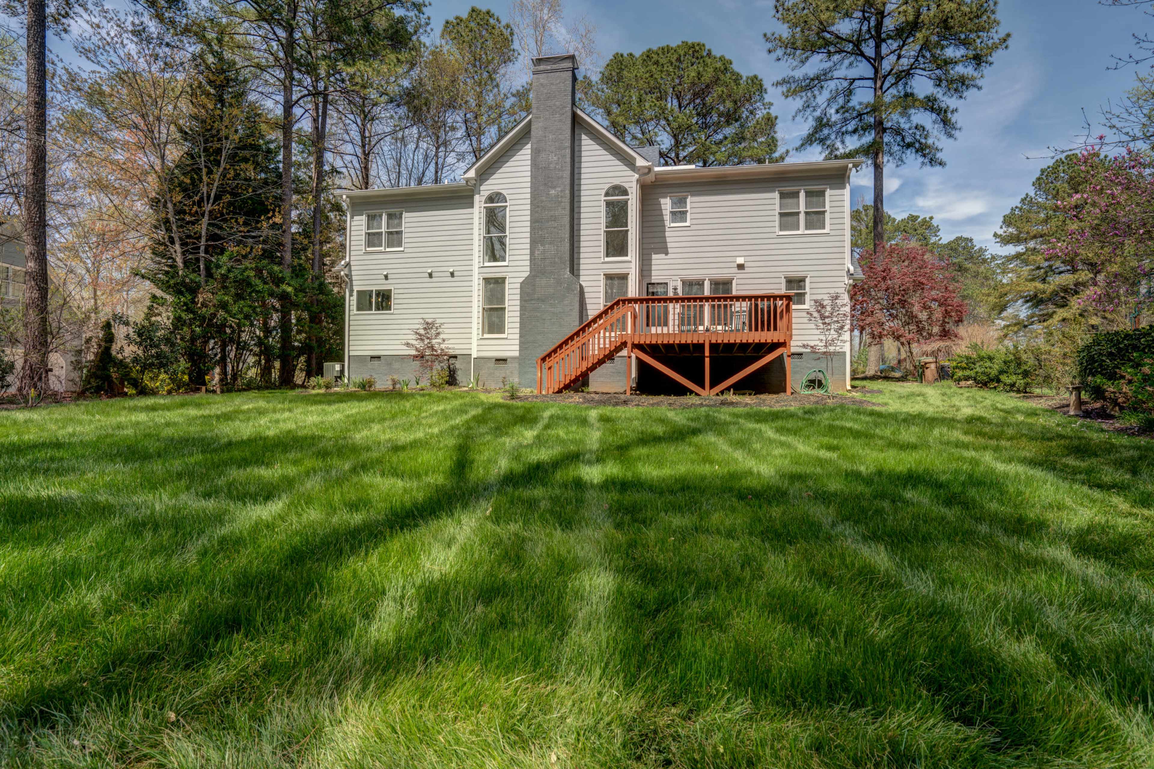 A two-story house with a gray chimney, a wooden deck in the back, and a large grassy yard surrounded by trees.