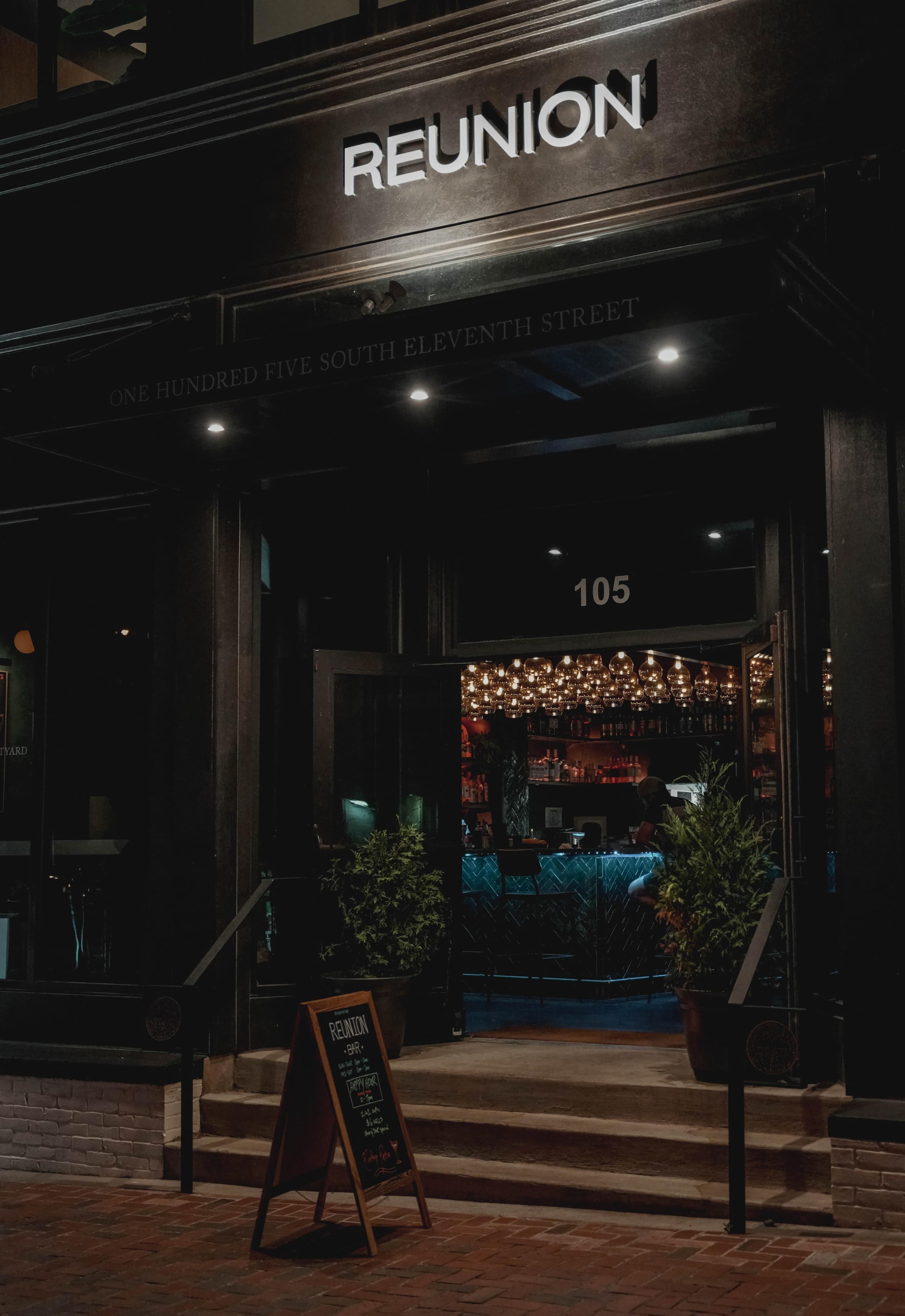 The entrance of a bar named "REUNION" is illuminated at night, featuring a prominent sign and decorative lighting inside.