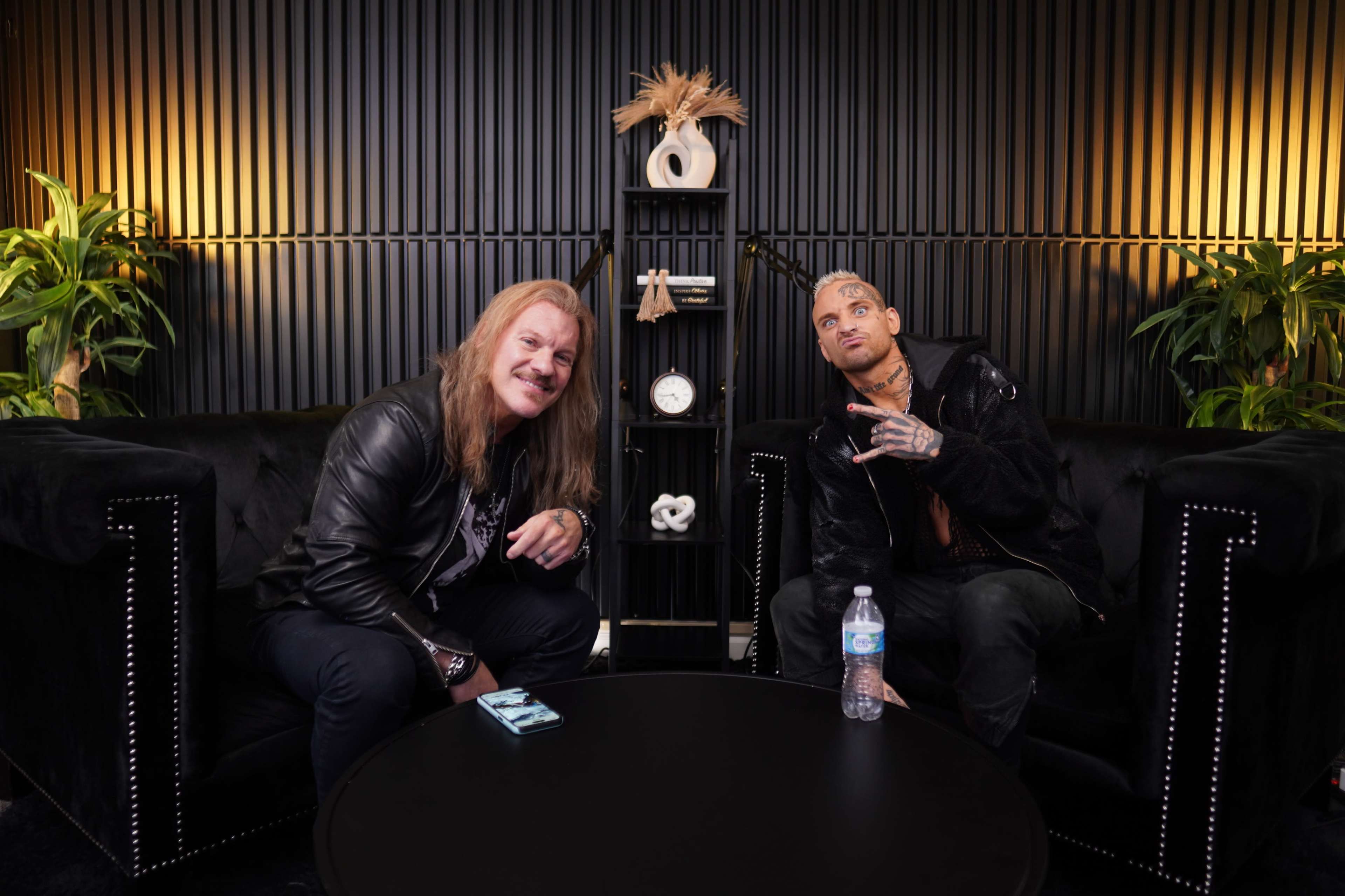 Two men are seated in a modern lounge area with black furniture, surrounded by decorative plants and a shelf displaying items.