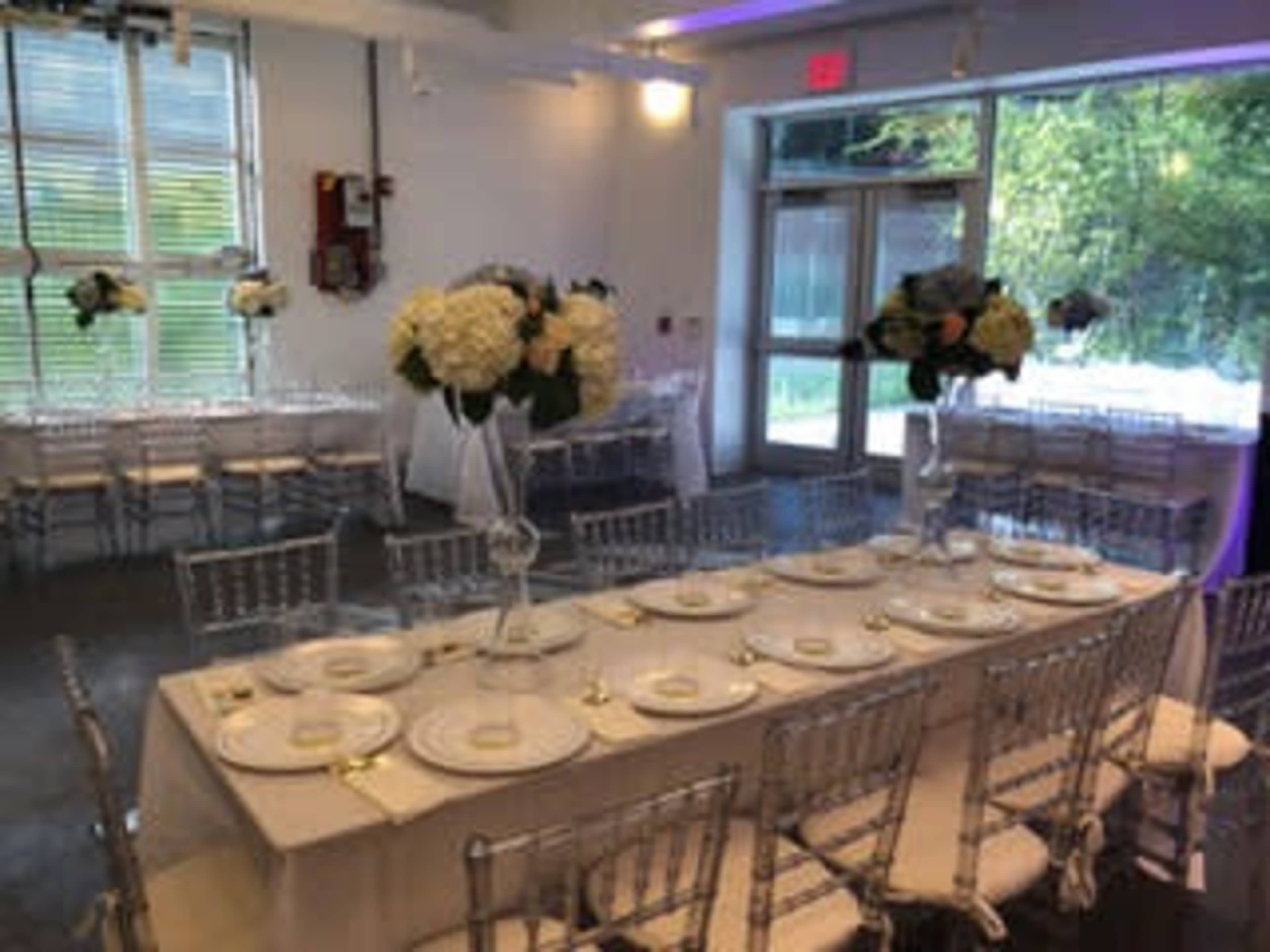 The image shows an elegantly arranged indoor event space with a long dining table set for a formal occasion, adorned with floral centerpieces and surrounded by chairs.