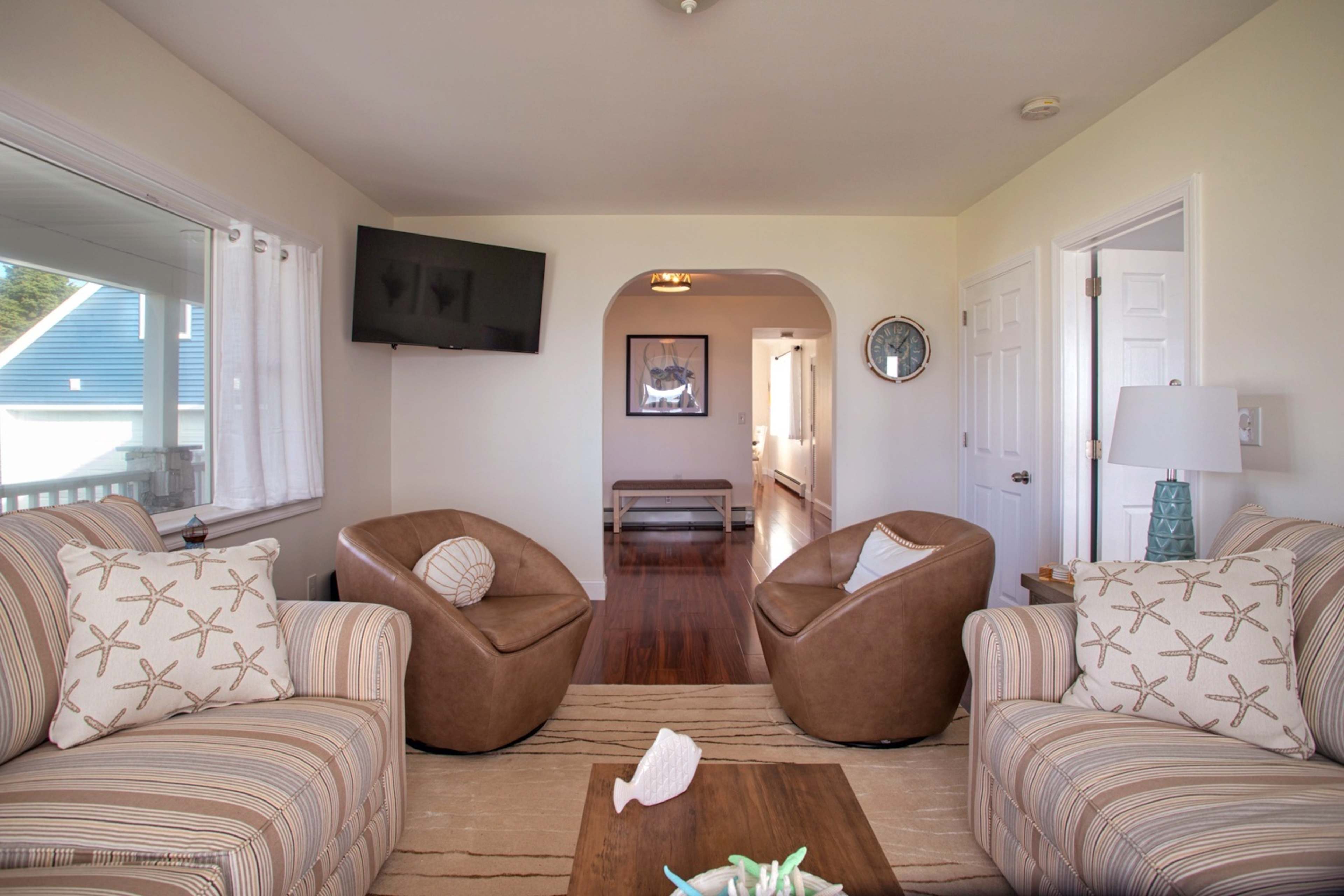 A cozy living room features two brown armchairs and a sofa adorned with starfish patterns, with a flat-screen TV mounted on the wall and a hallway leading to other areas.