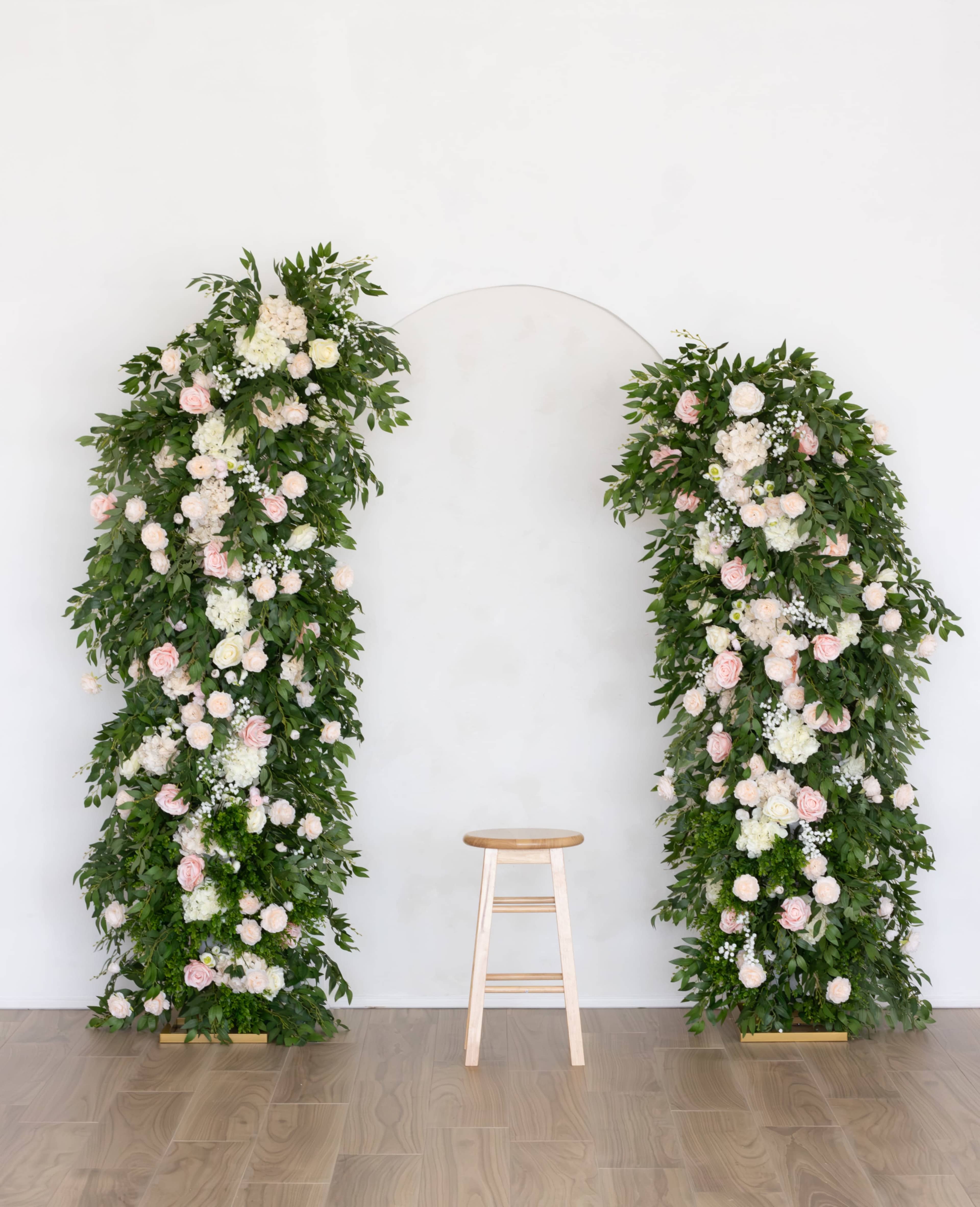 The image features a minimalist floral arch adorned with greenery and white and pink flowers, flanking a simple wooden stool against a white wall.
